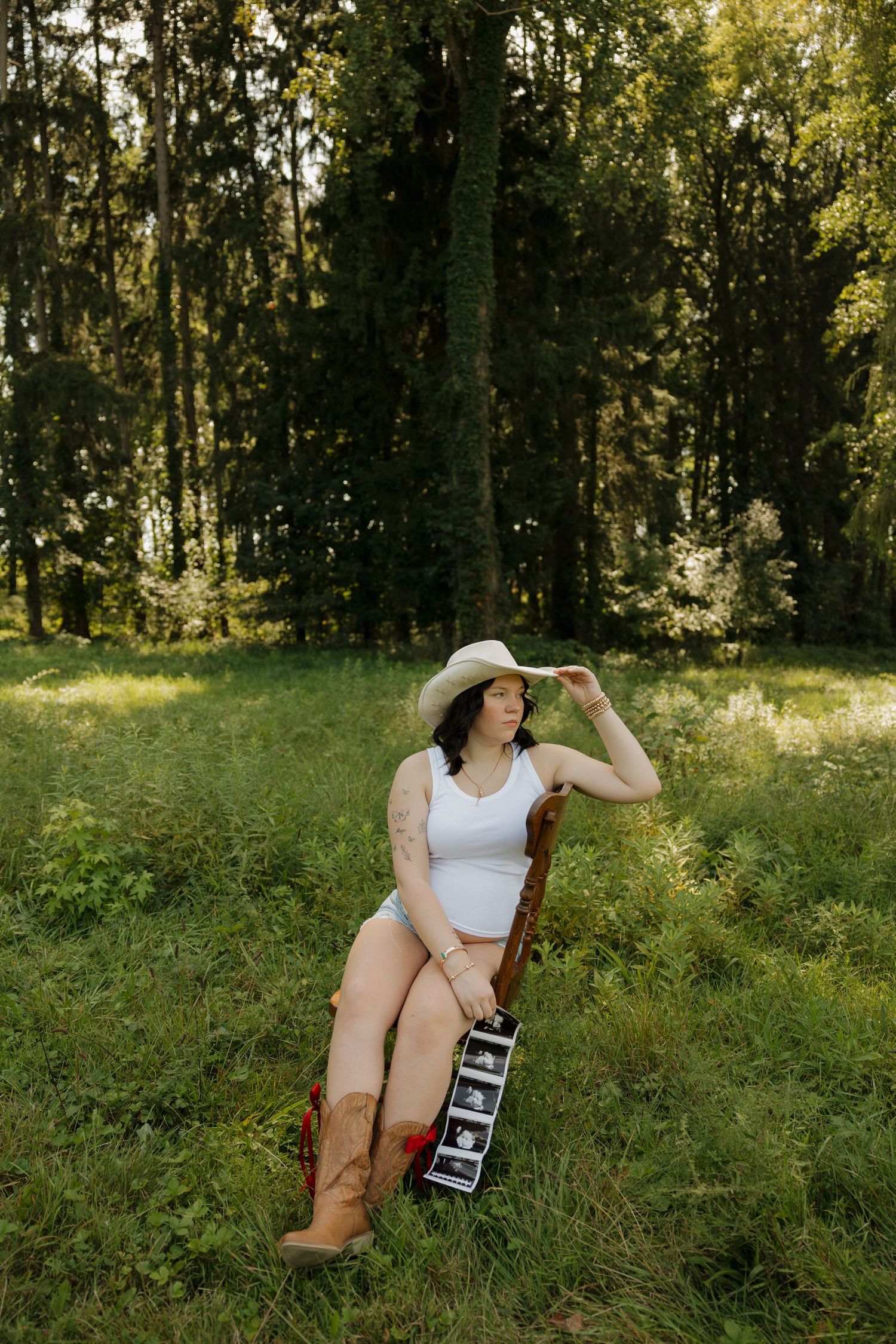 Pregnant woman in white bodysuit, cowboy hat, and boots sits in chair outdoors holding ultrasound photos in lush green forest.
