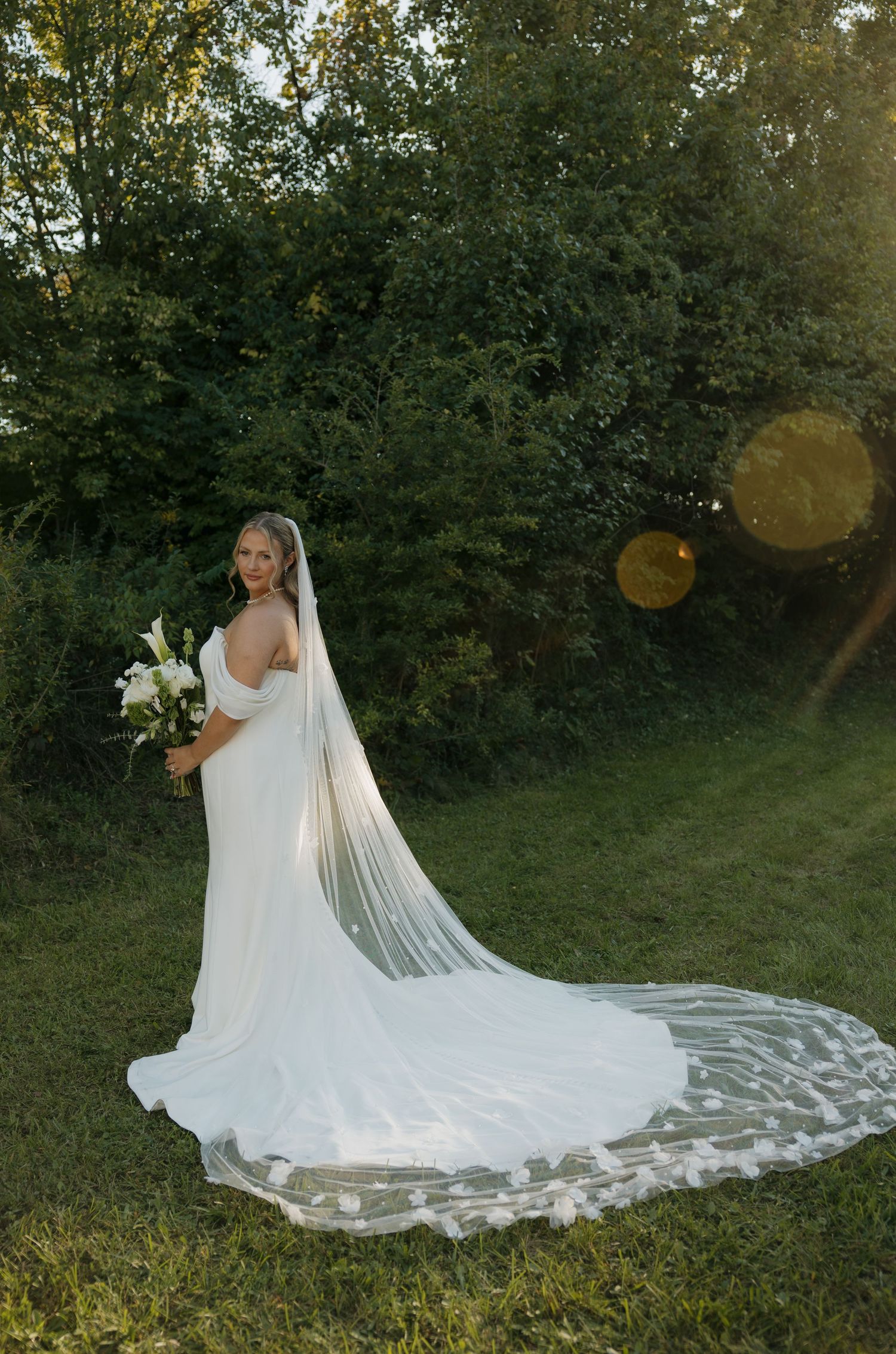 Bride in white gown with long lace veil holding bouquet, standing on green grass with trees in background.