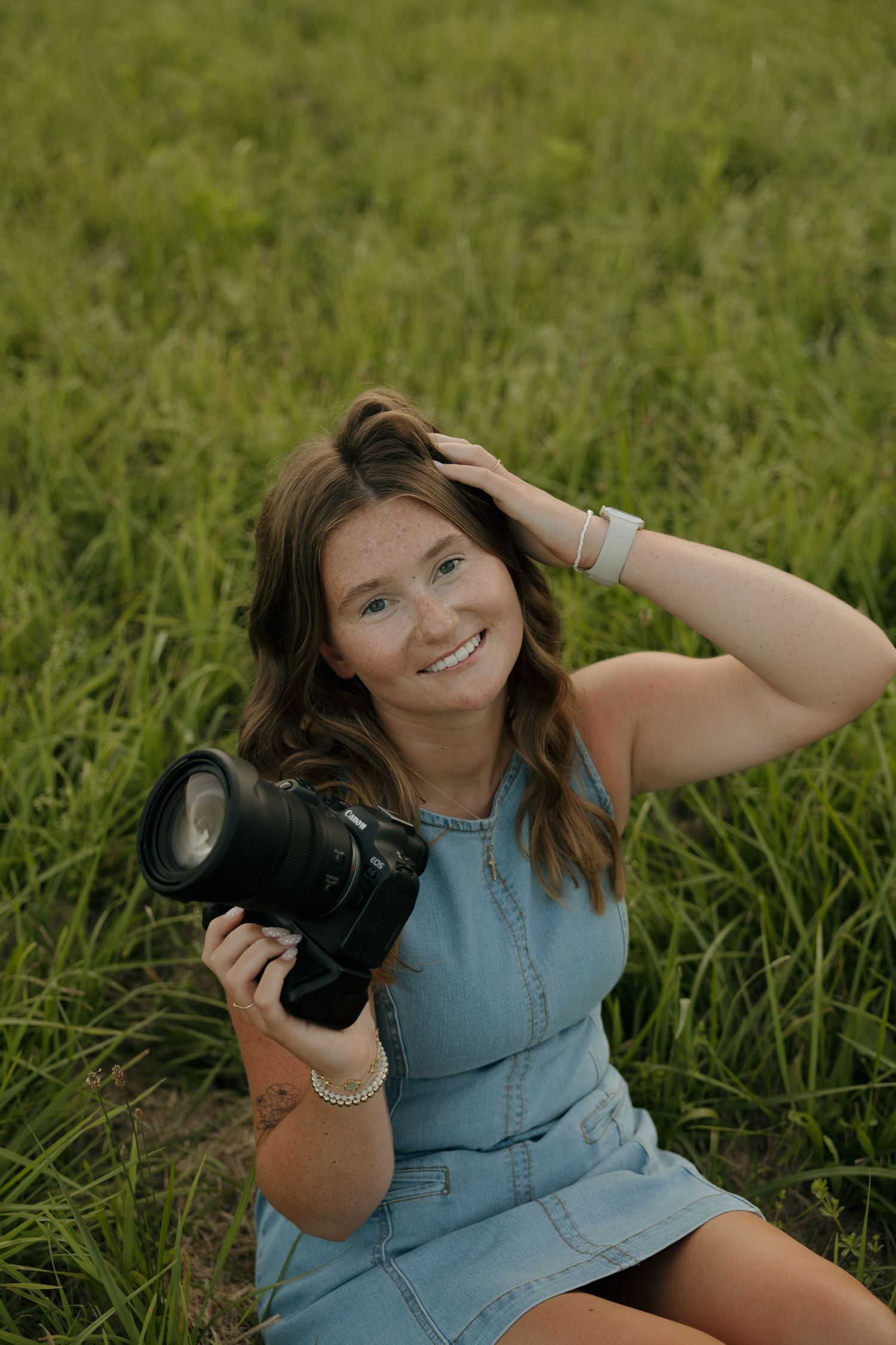 Young woman in denim dress sitting in green field, smiling and holding a professional DSLR camera.