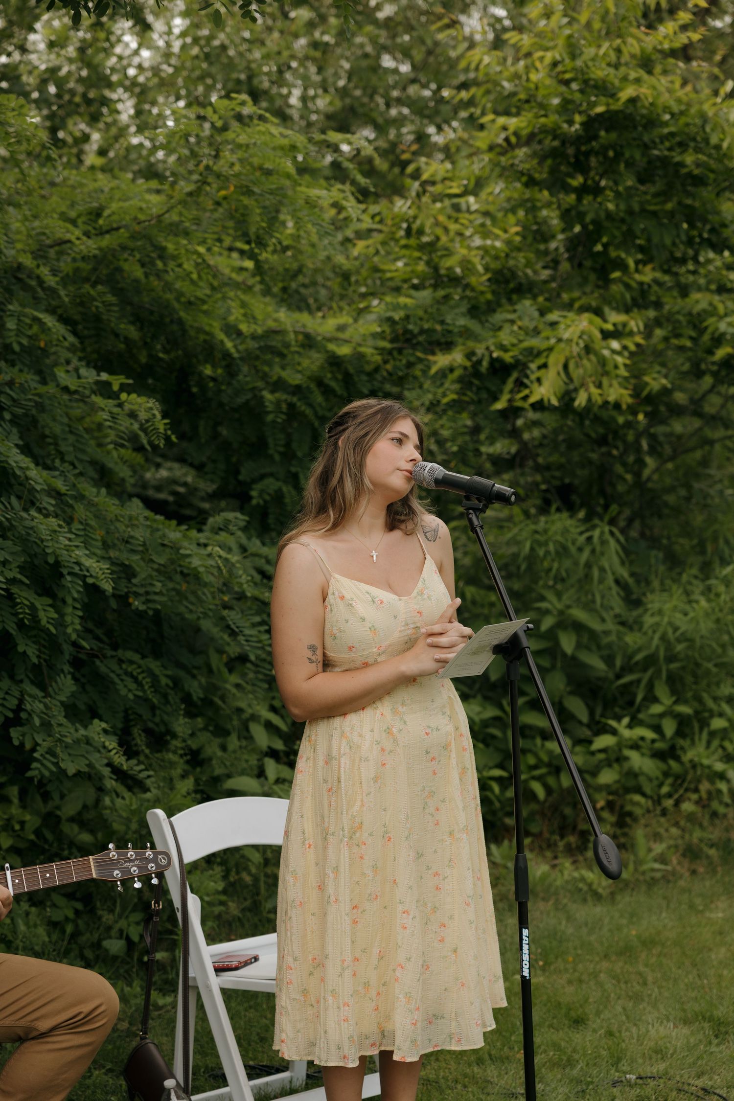 Woman in floral yellow dress speaking at outdoor microphone stand with lush green trees in background.