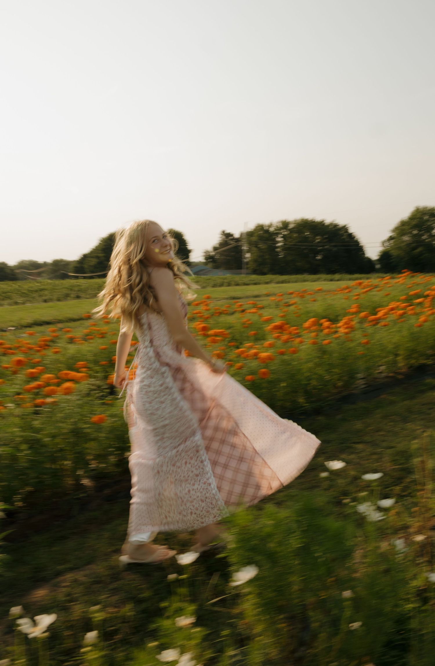A woman in a flowing pink patchwork dress twirls in a vibrant field of orange flowers on a sunny day.