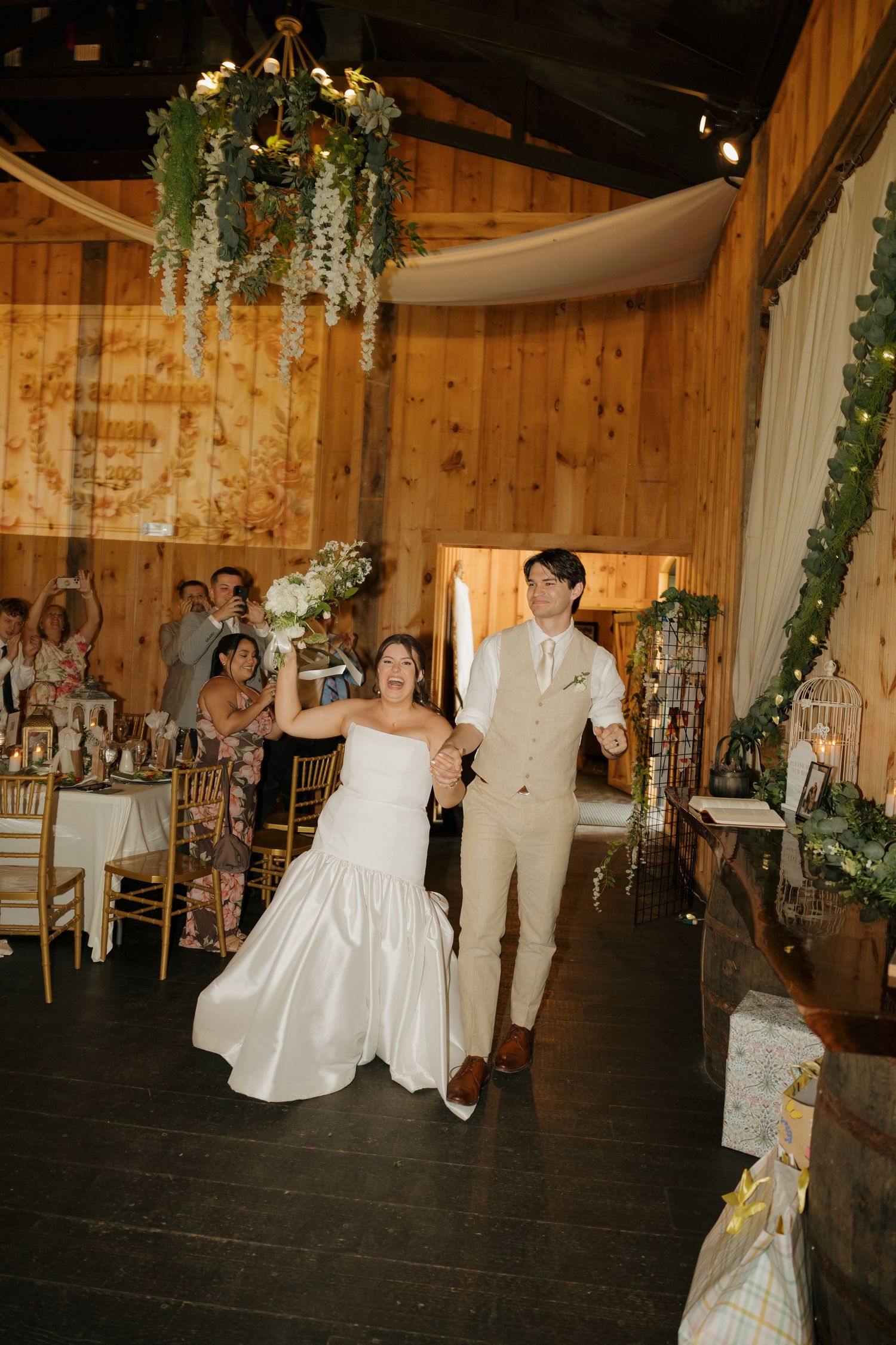 Bride and groom make grand entrance into rustic barn wedding reception, bride holding bouquet aloft, guests cheering.