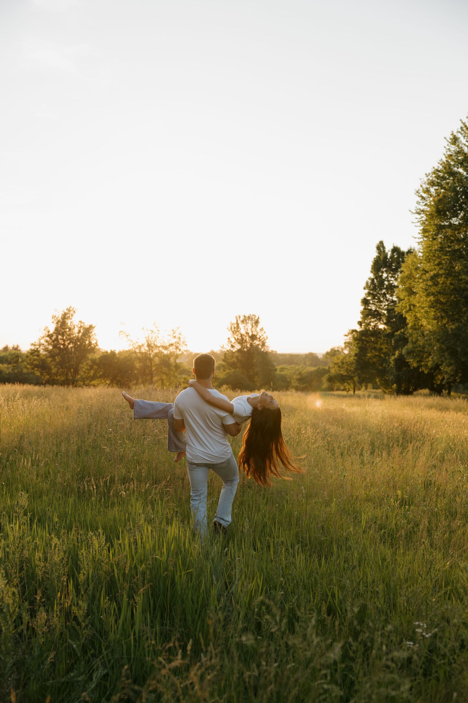 Couple dancing in a golden sunset field, man lifting woman in white outfits, surrounded by lush green grass and trees.