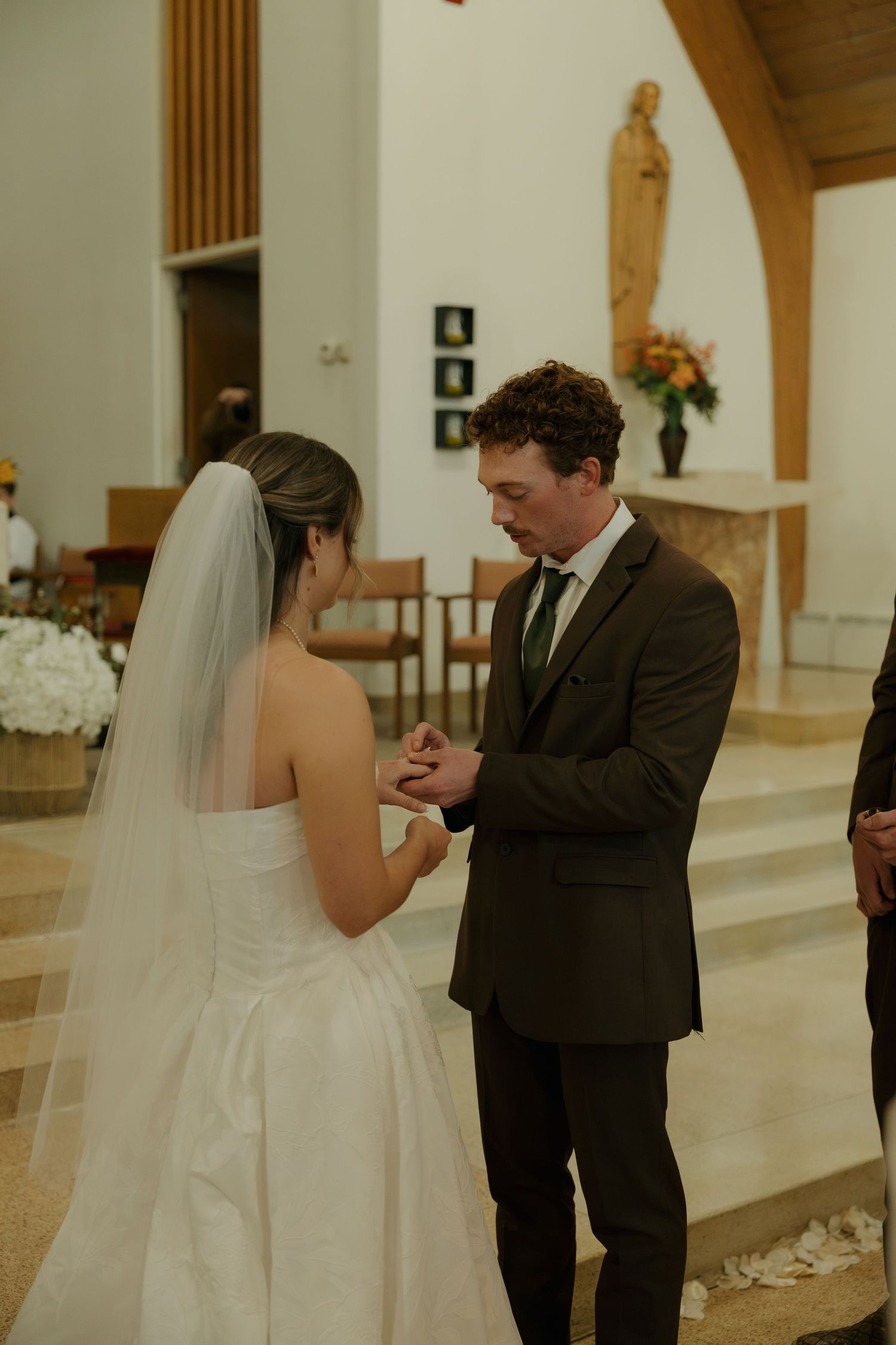 A groom in a brown suit places a ring on his bride's finger during a church wedding ceremony, as she stands in a white gown and veil.