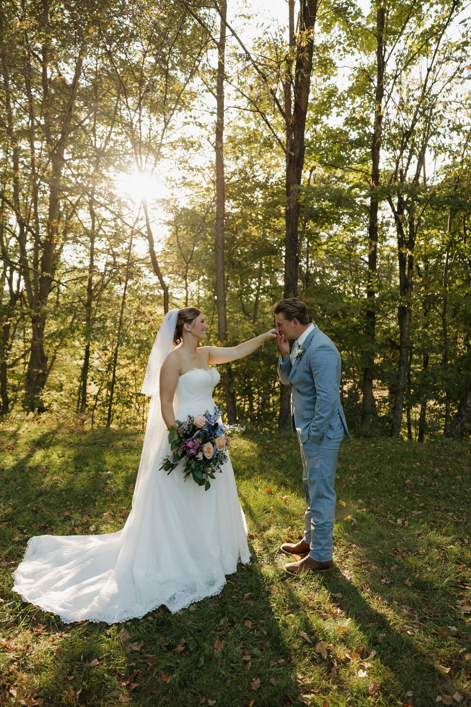 Groom in a light blue suit kisses the hand of a bride in a white gown holding a bouquet in a sunlit forest clearing.