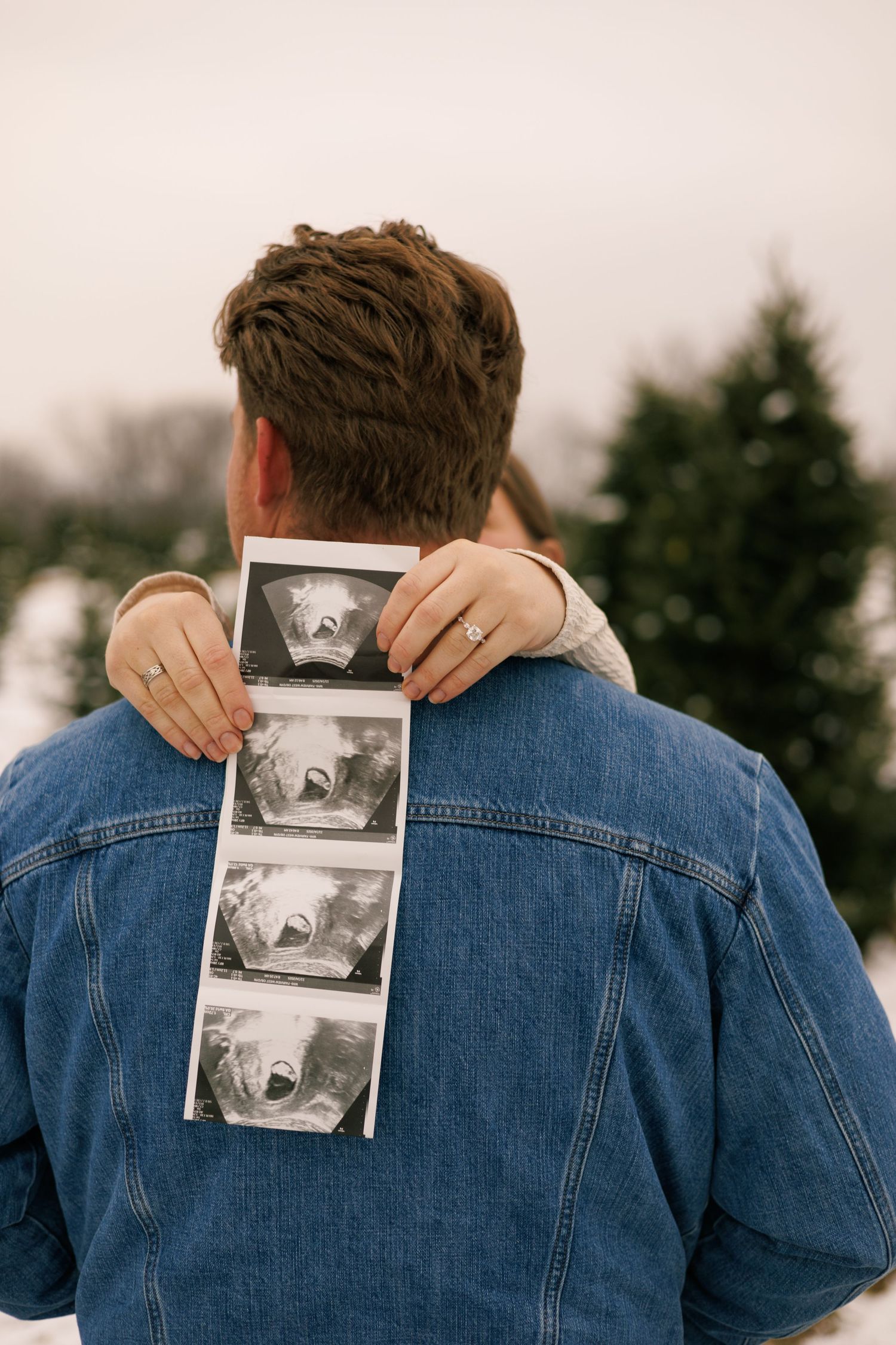 A person in a denim jacket holds up a sonogram photo against a winter backdrop.