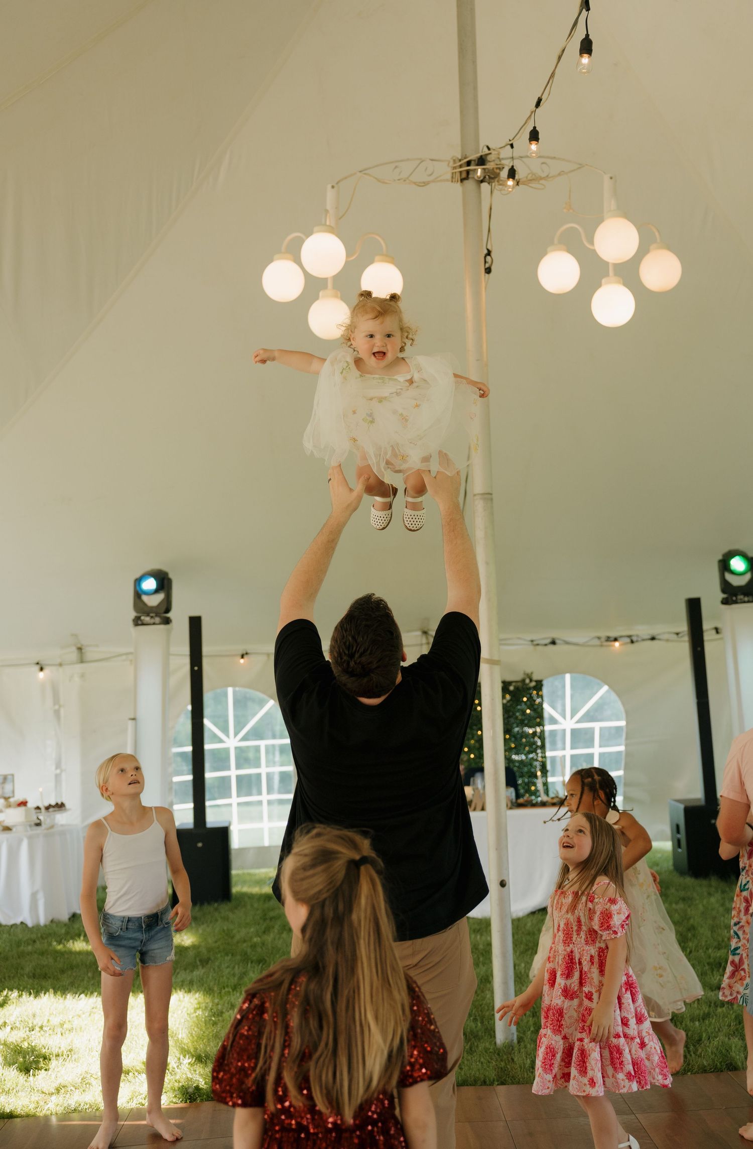 Joyful man tosses laughing toddler in white dress high in air inside decorated wedding reception tent.