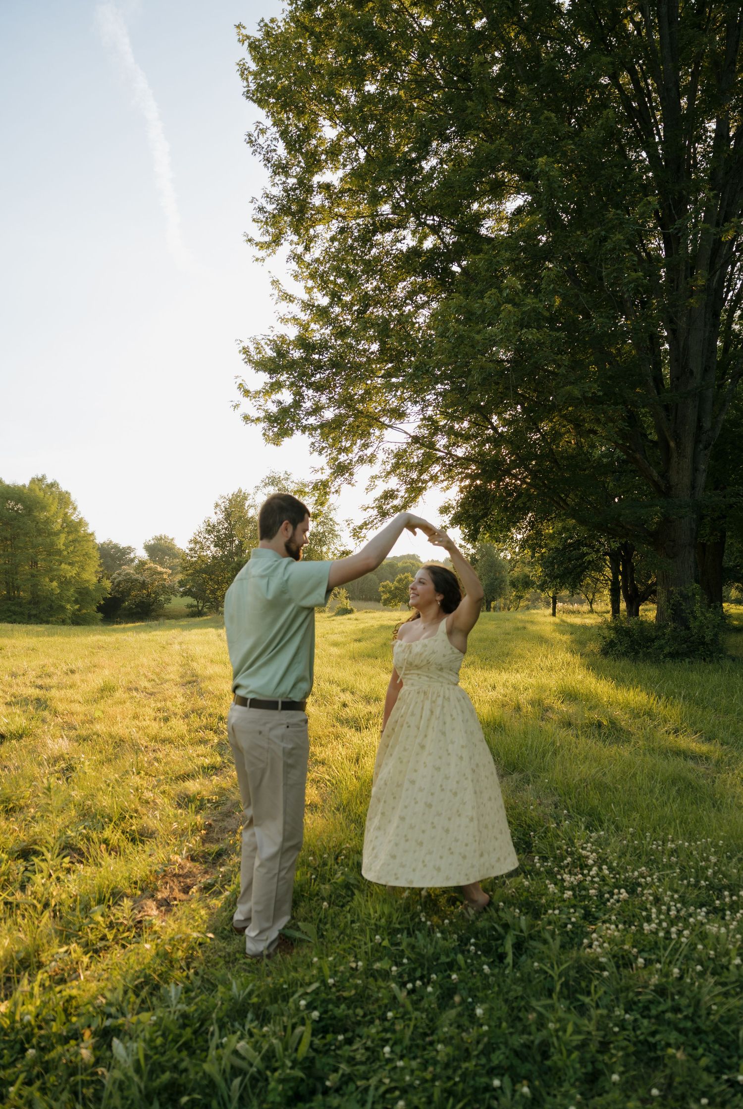 Couple dancing together in a sunny green meadow surrounded by tall trees during golden hour.