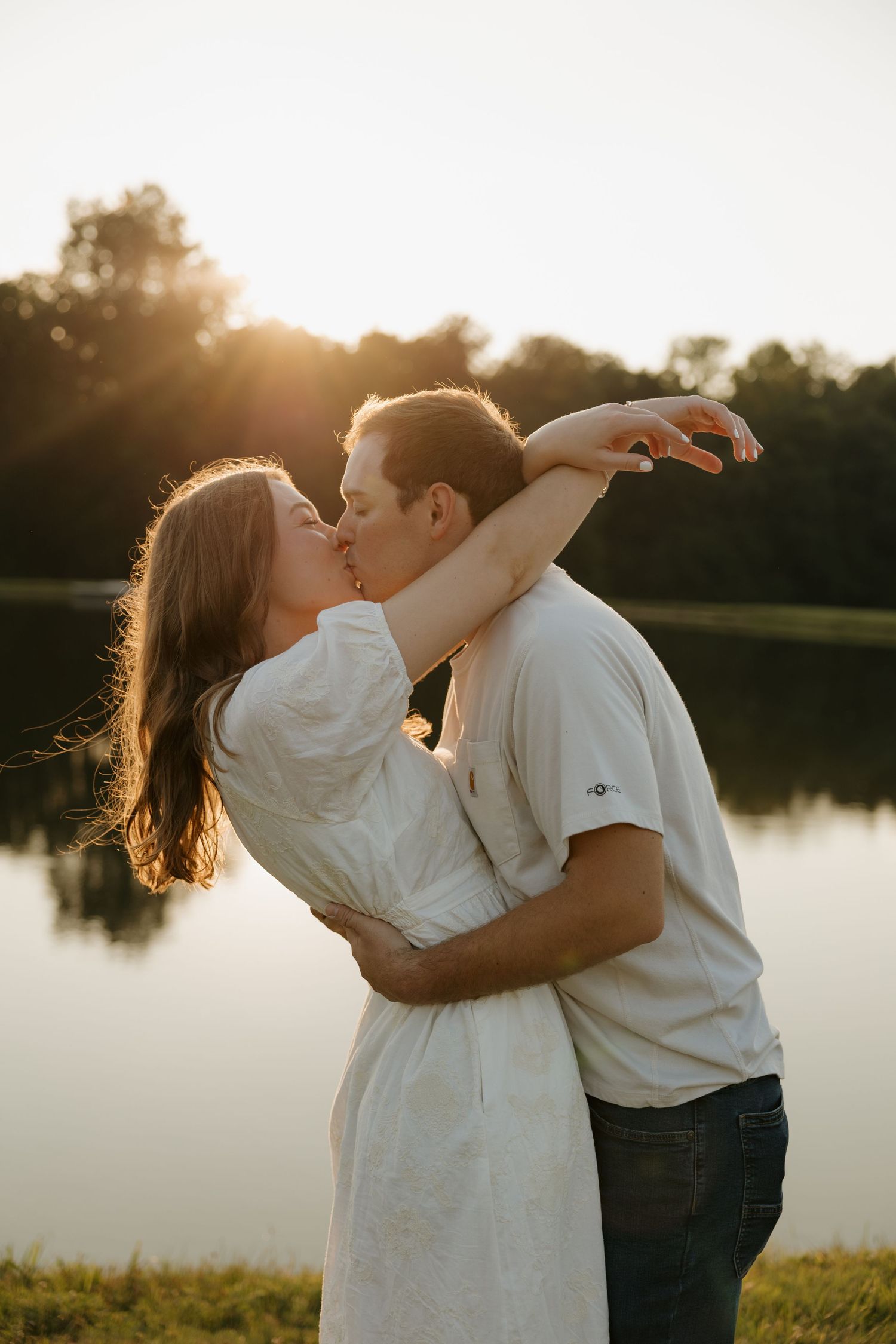 Couple sharing a romantic kiss at golden hour by a reflective lake, woman in white dress with arms around mans neck.
