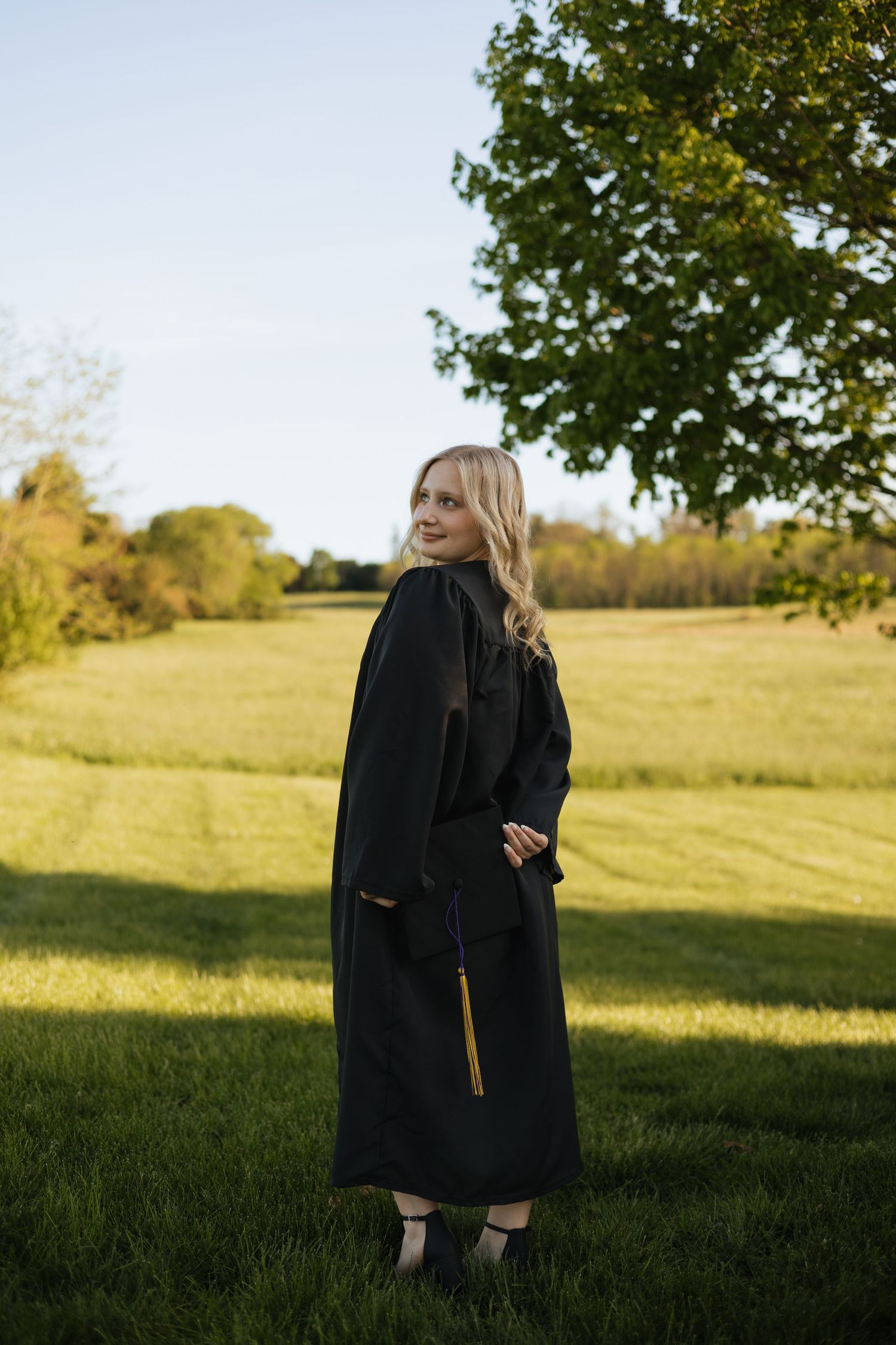 Blonde graduate in black cap and gown standing in green field with golden sunlight.