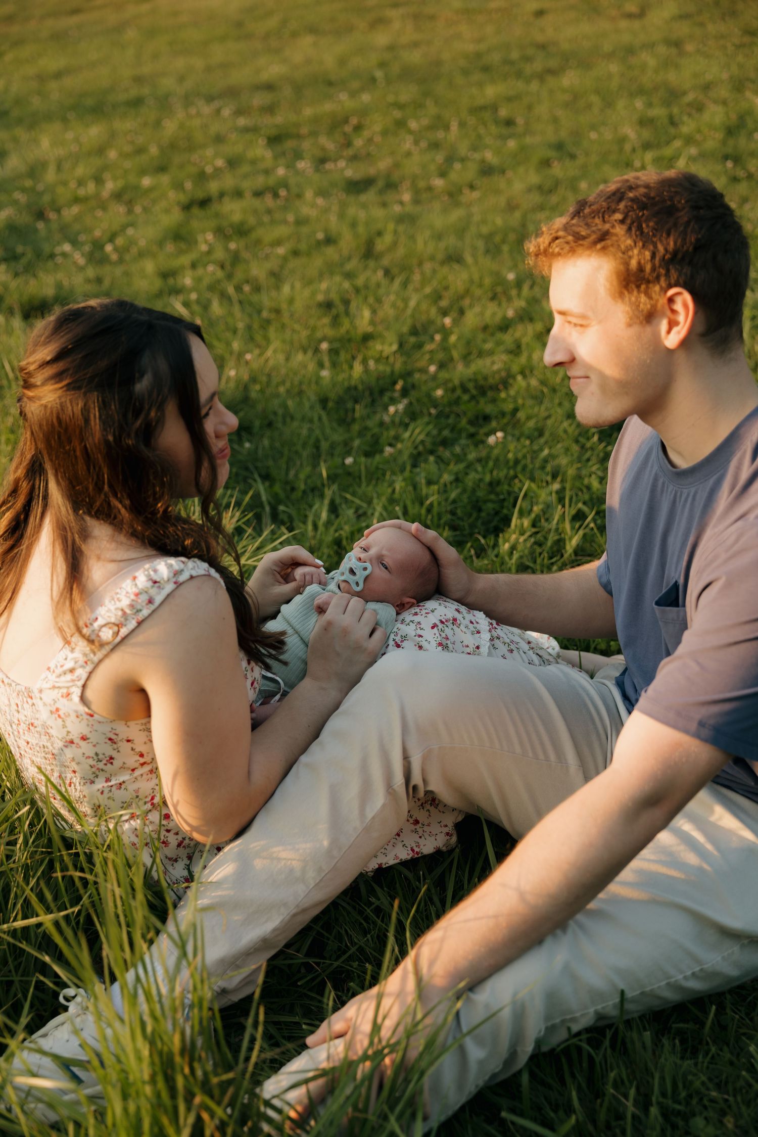 Young couple sitting in grass at sunset, lovingly attending to their newborn baby.