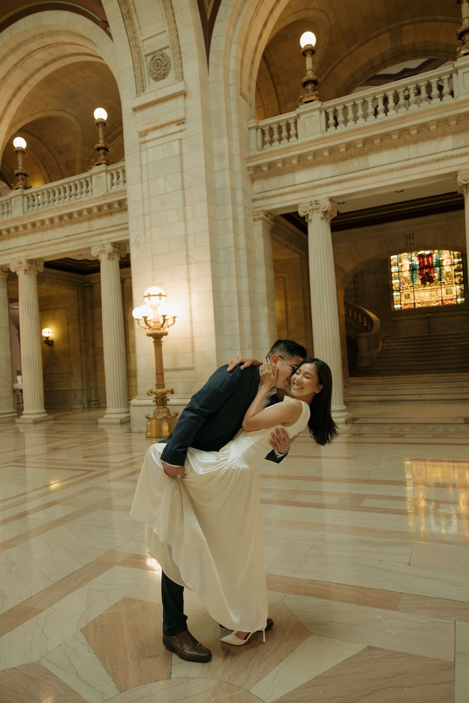 Couple sharing a romantic dip kiss in an elegant marble hall with grand columns and ornate architecture.