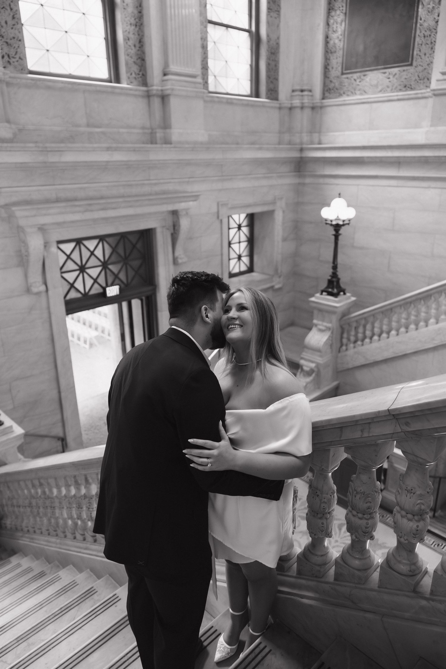 Black and white photo of a couple embracing on grand marble staircase in elegant building, bride in white dress.