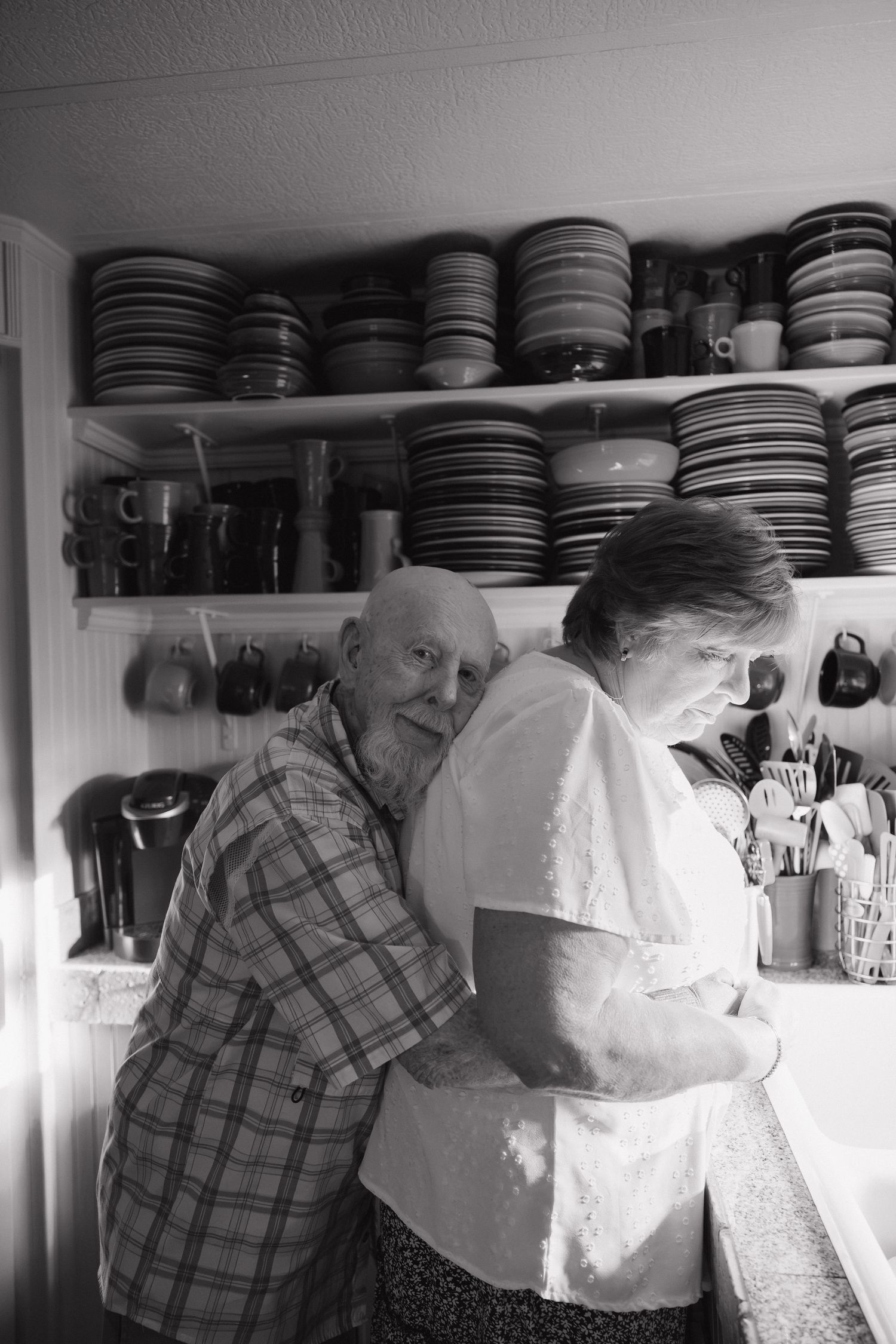 Elderly couple sharing a tender embrace in a black and white kitchen photo with shelves of dishes.
