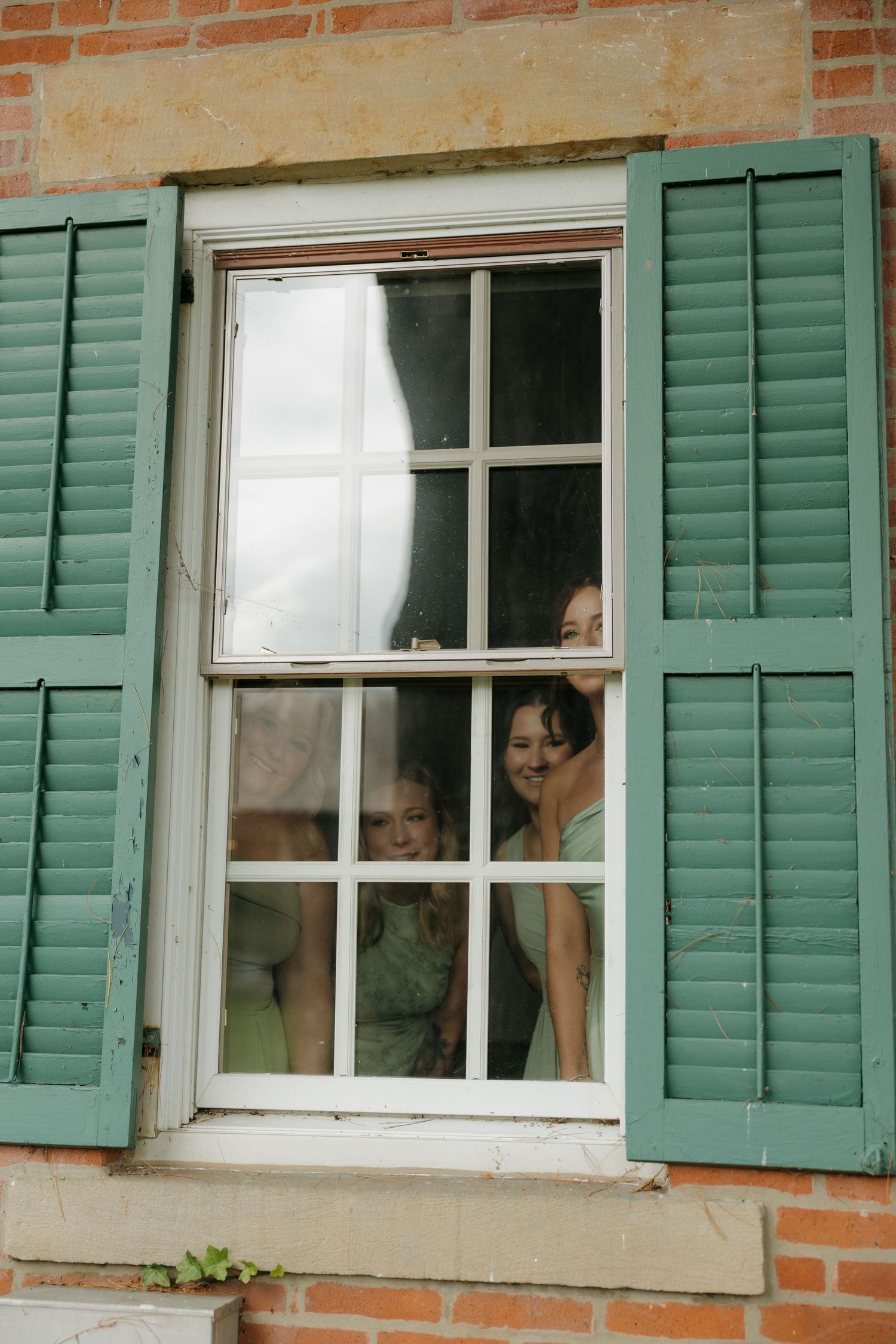 Two women peering through a white-framed window with green shutters on a brick building.