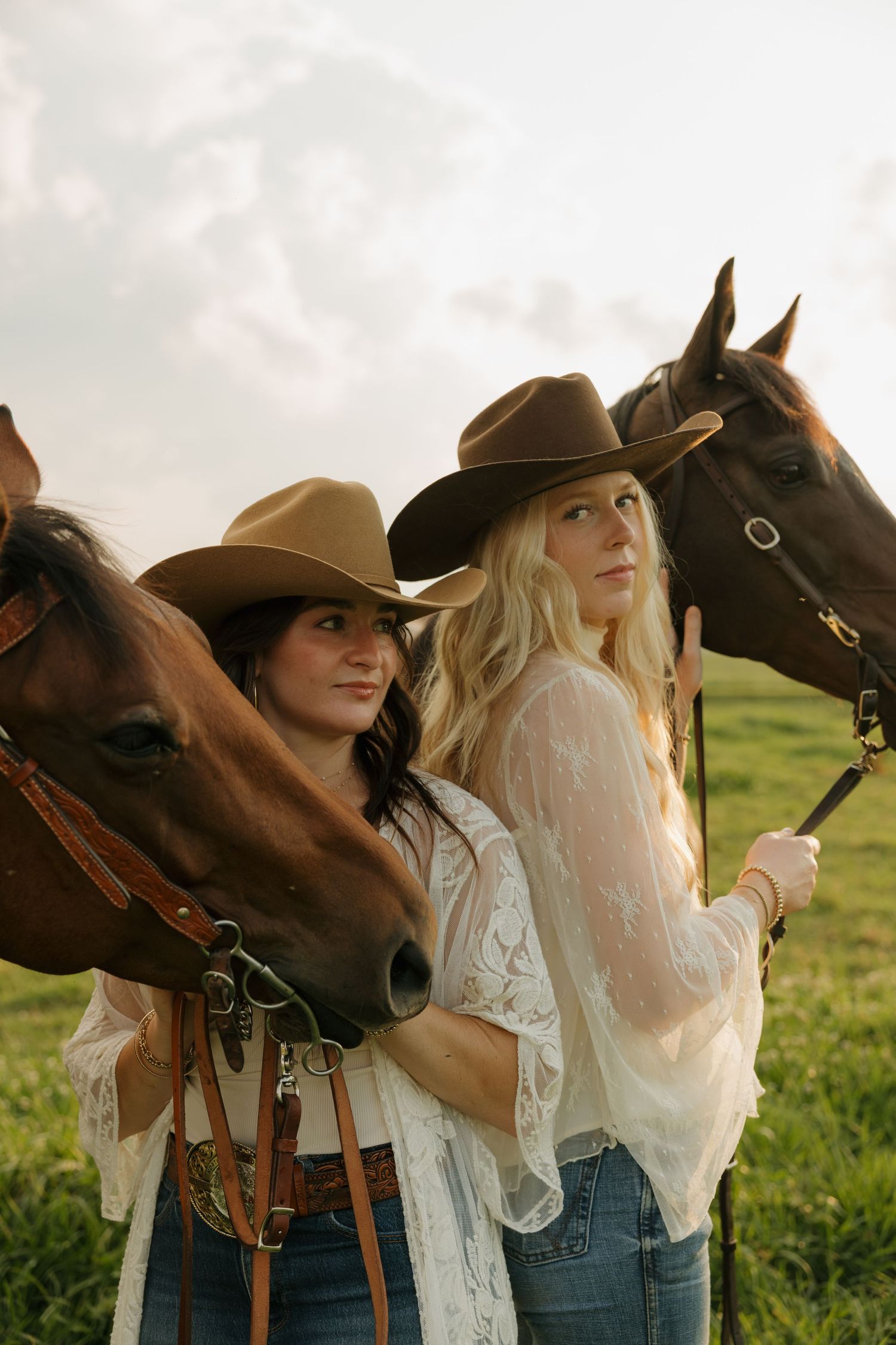Two women in cowboy hats pose with brown horses in a green field, wearing white lace tops and jeans.