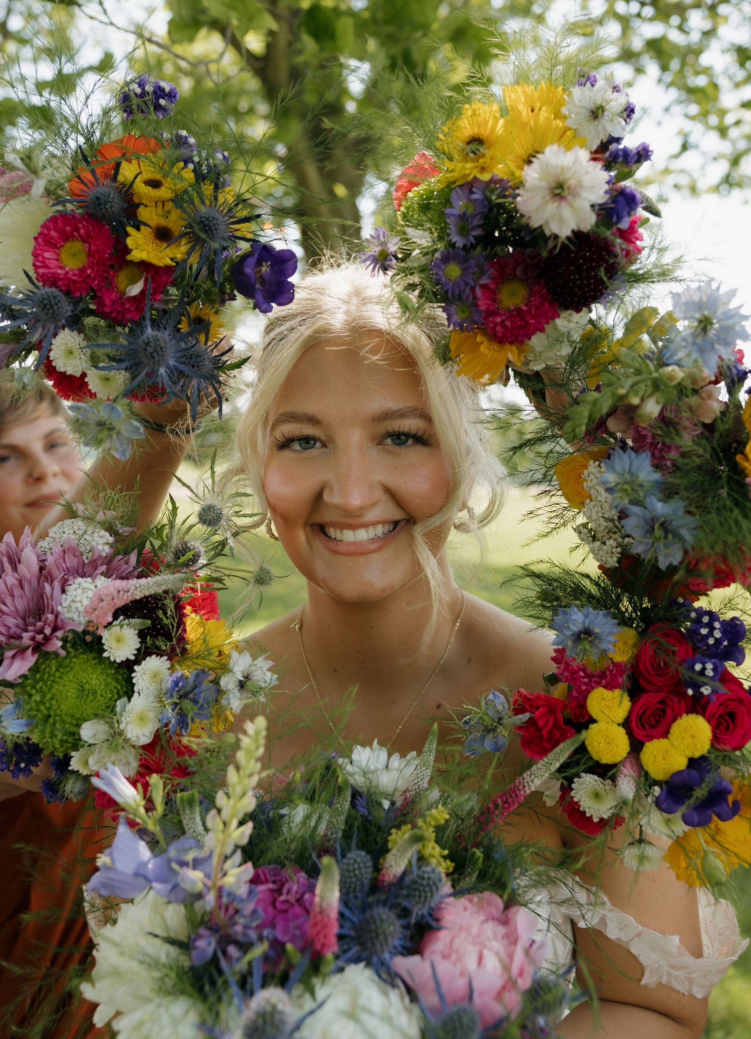 Smiling bride framed by colorful wildflower bouquets with mixed blooms including daisies, thistles, and sunflowers outdoors.