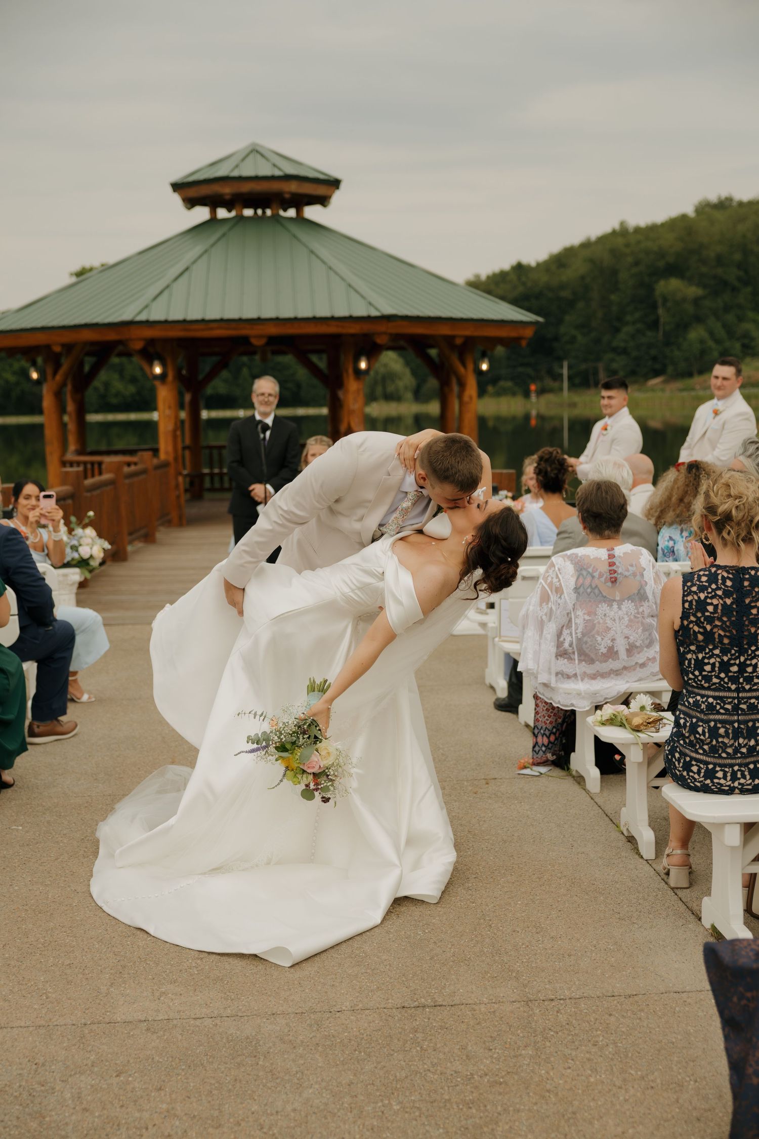 Bride and groom share a romantic dip kiss on a wooden dock with a gazebo backdrop during outdoor wedding ceremony.