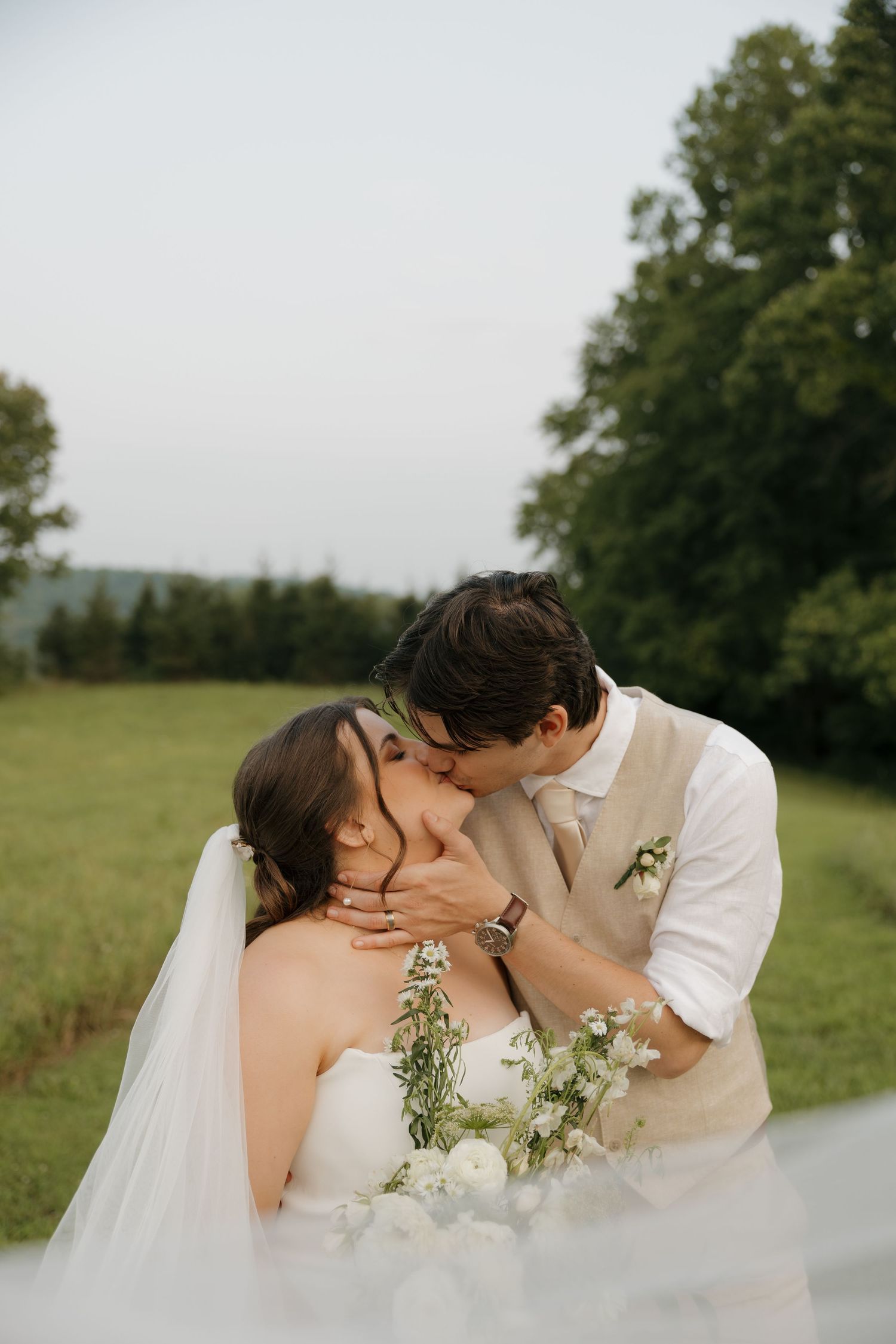 Groom kissing bride on the cheek in a green field, bride holding white bouquet with veil flowing in the breeze.