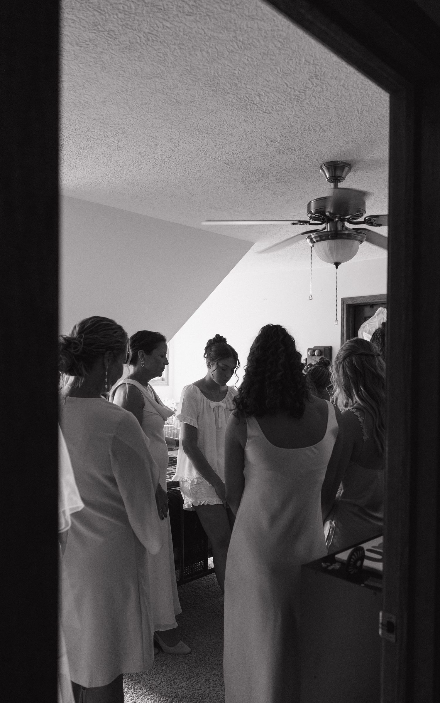 Black and white photo of bridesmaids in white dresses preparing together in a room with a ceiling fan.