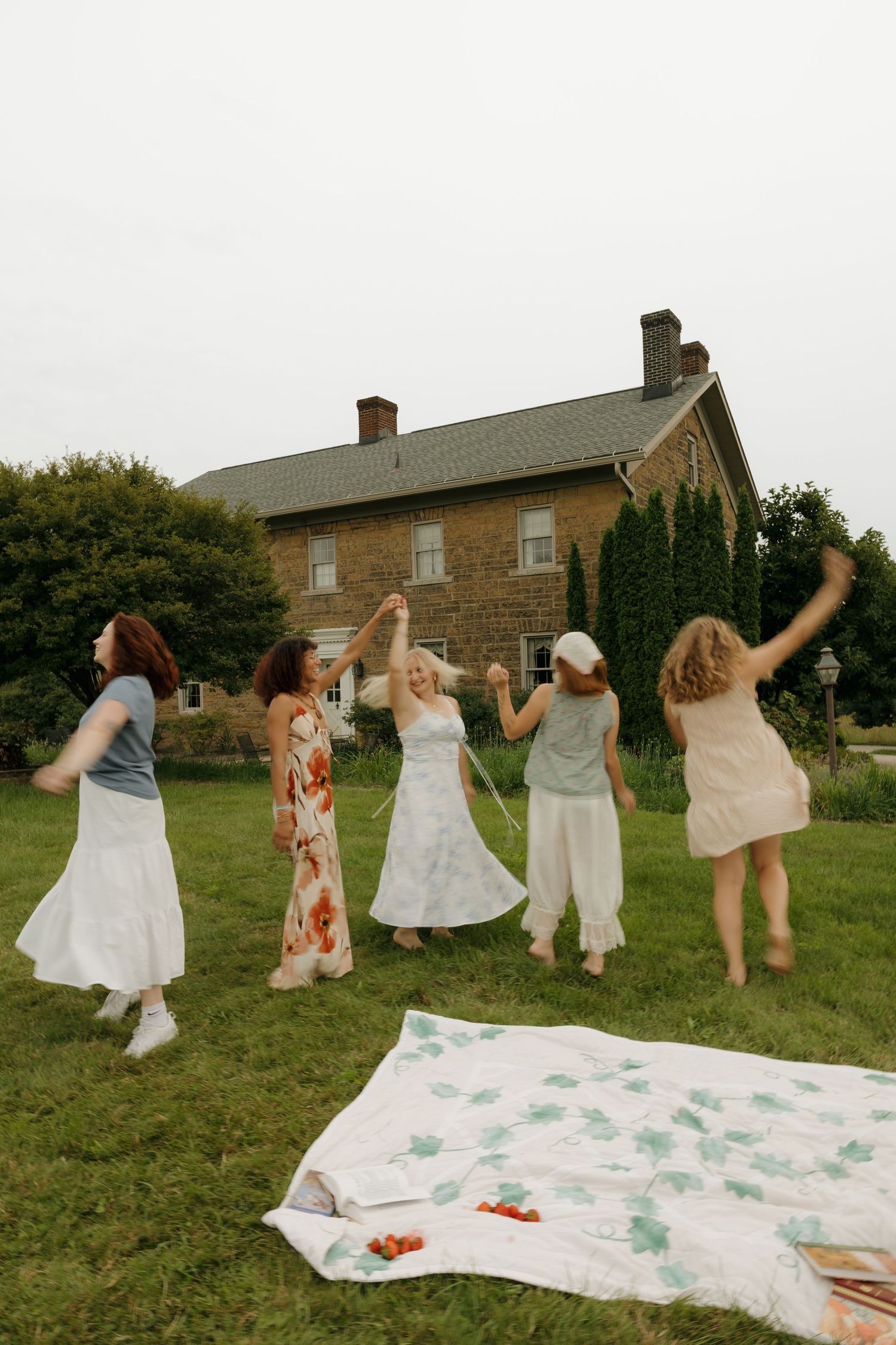 Five women dancing joyfully on green grass near a picnic blanket with a stone farmhouse in the background.
