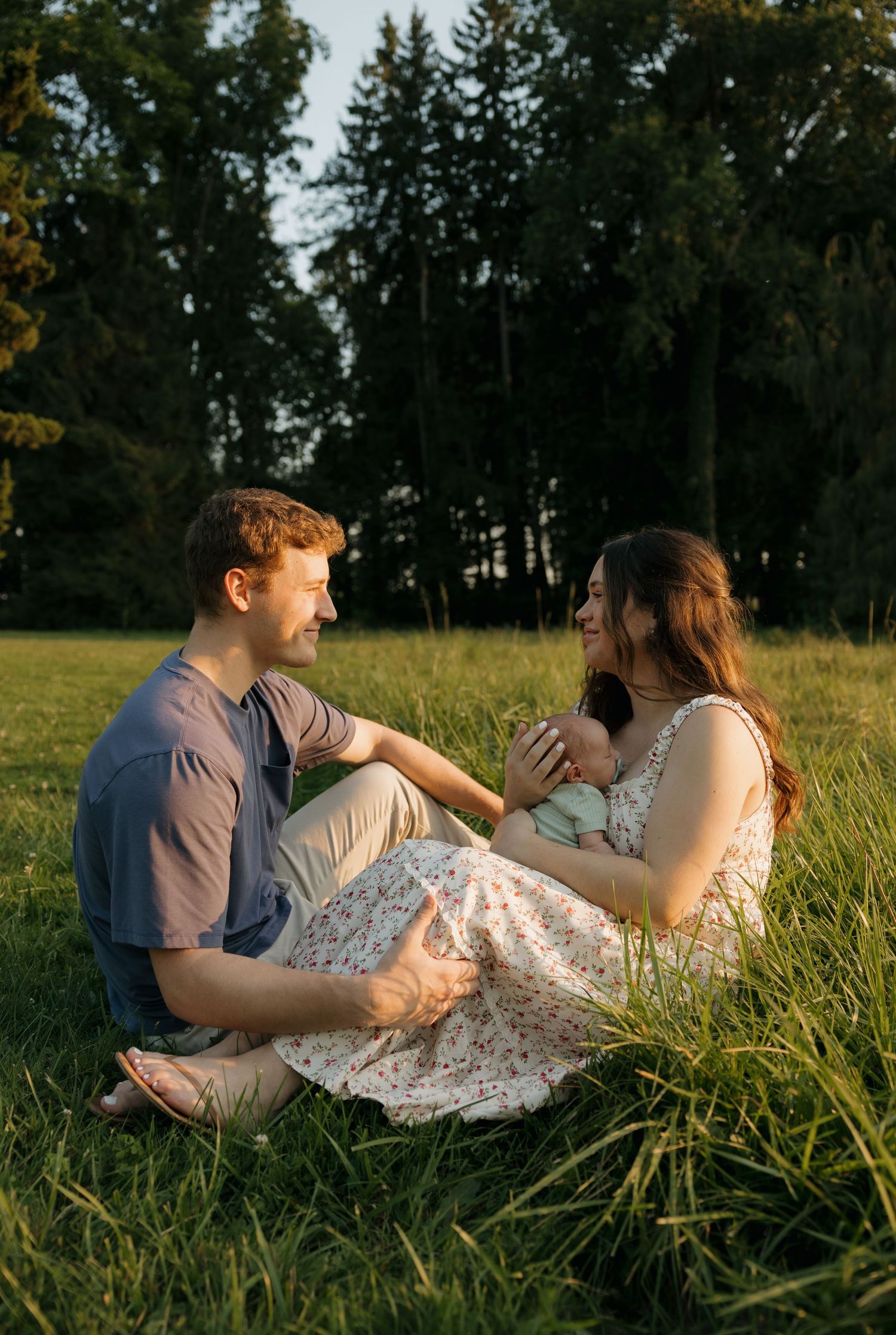 Young couple sits in lush grass at sunset, smiling at each other while cradling their newborn baby.