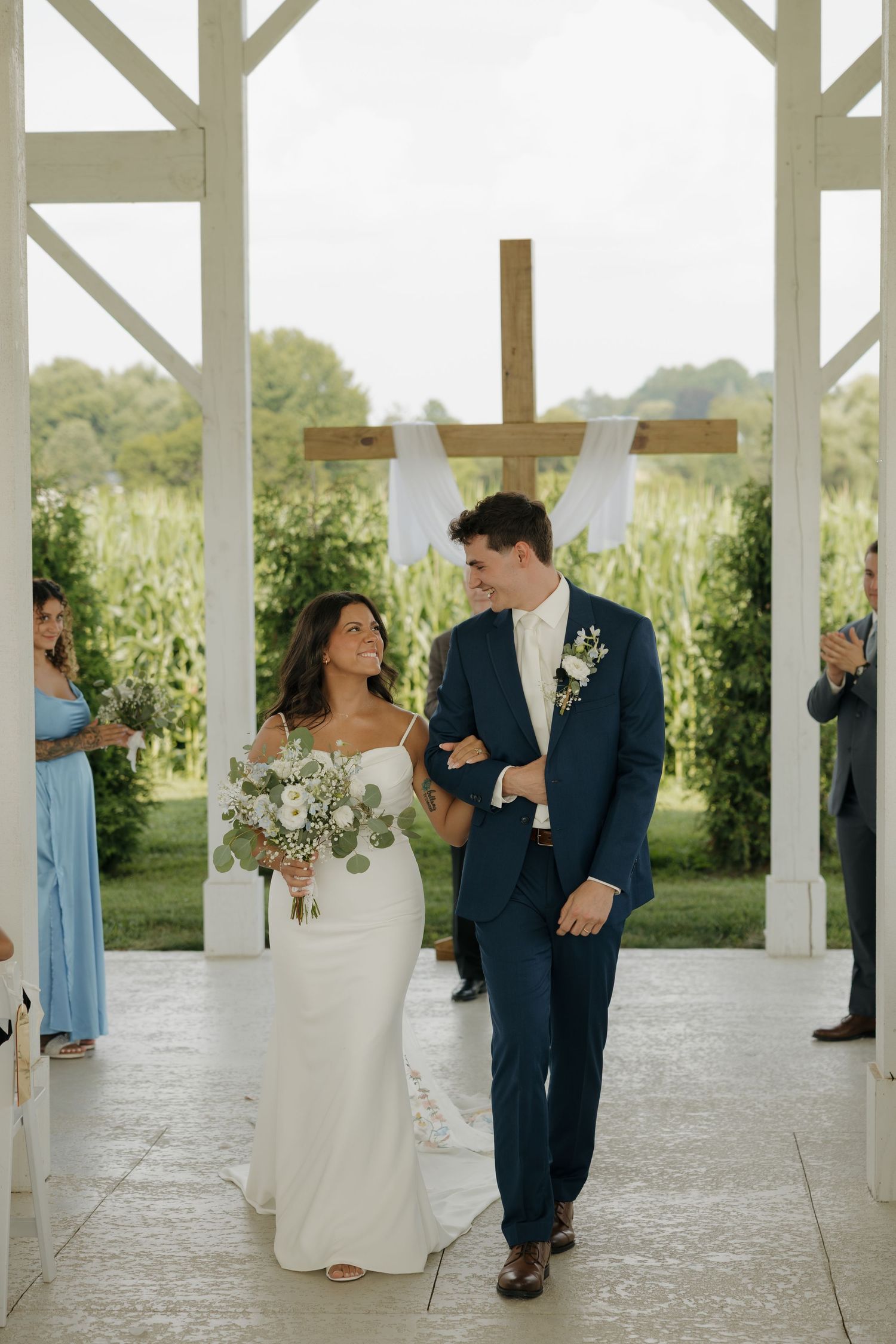 Newlyweds walk back down the aisle smiling after their ceremony at a white outdoor chapel.