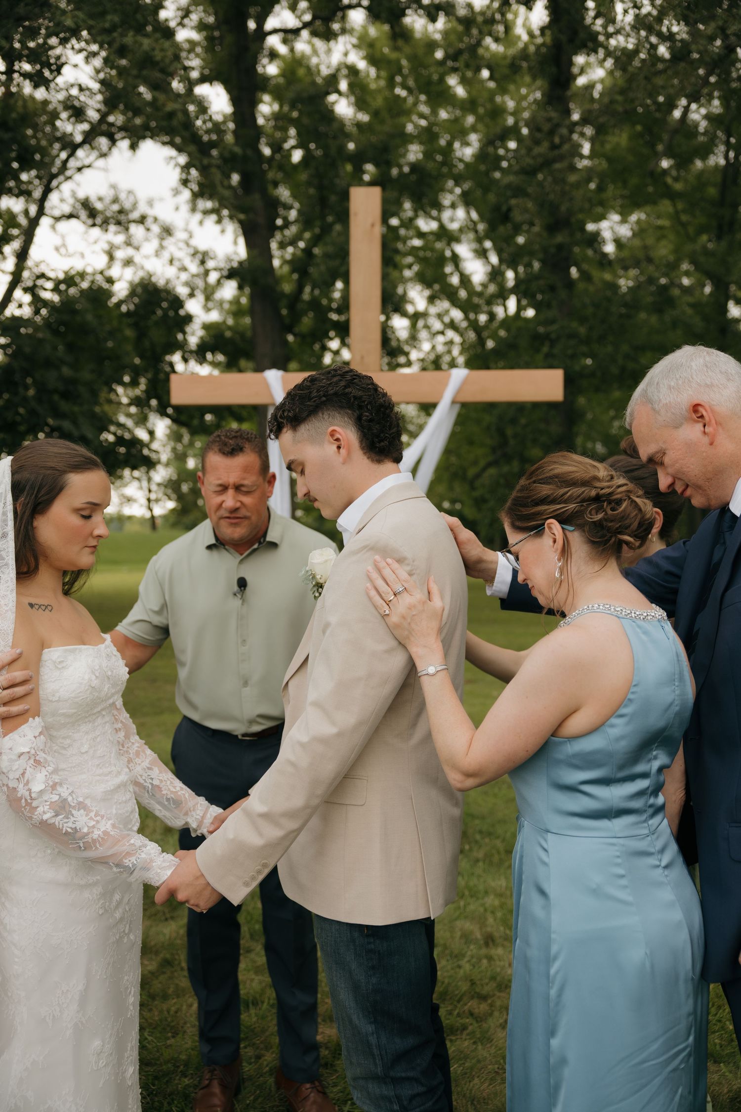 Wedding couple praying with family outdoors near a wooden cross, bride in lace gown and groom in beige suit.