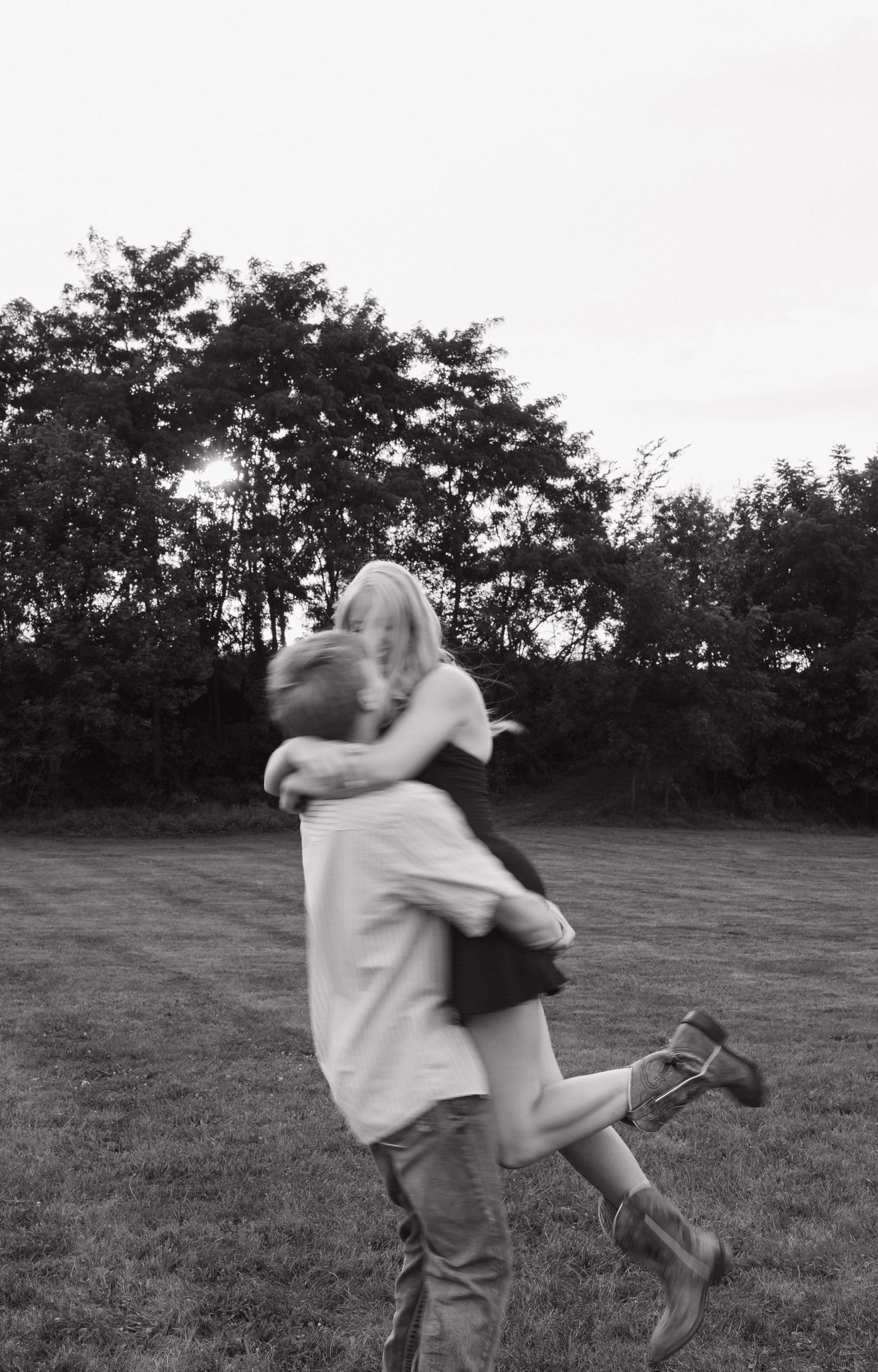 Black and white photo of a couple embracing and spinning joyfully on a grassy field with trees in the background.
