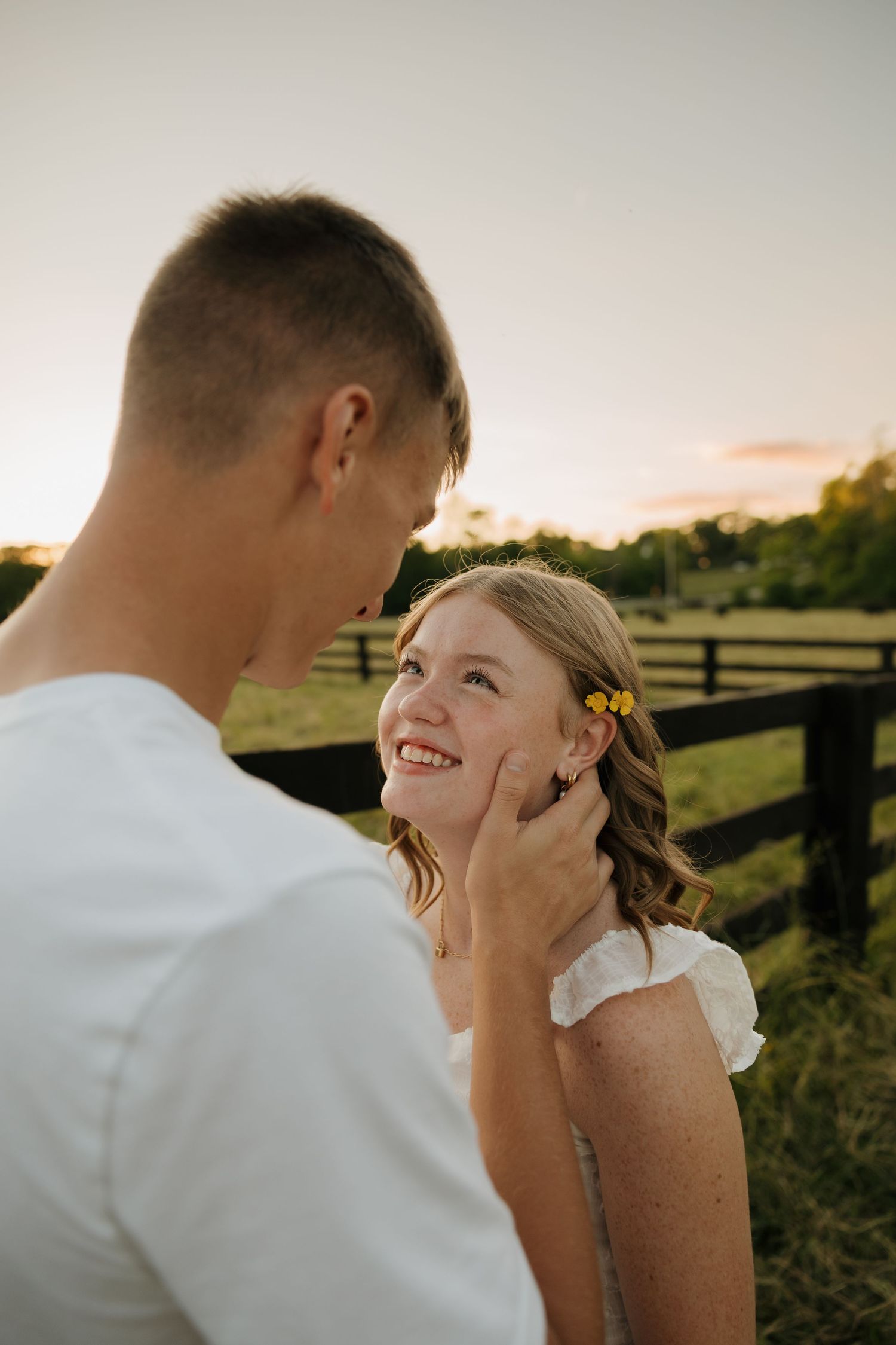 Young couple sharing a tender moment at sunset near a rustic fence, girl smiling with yellow flowers in her hair.