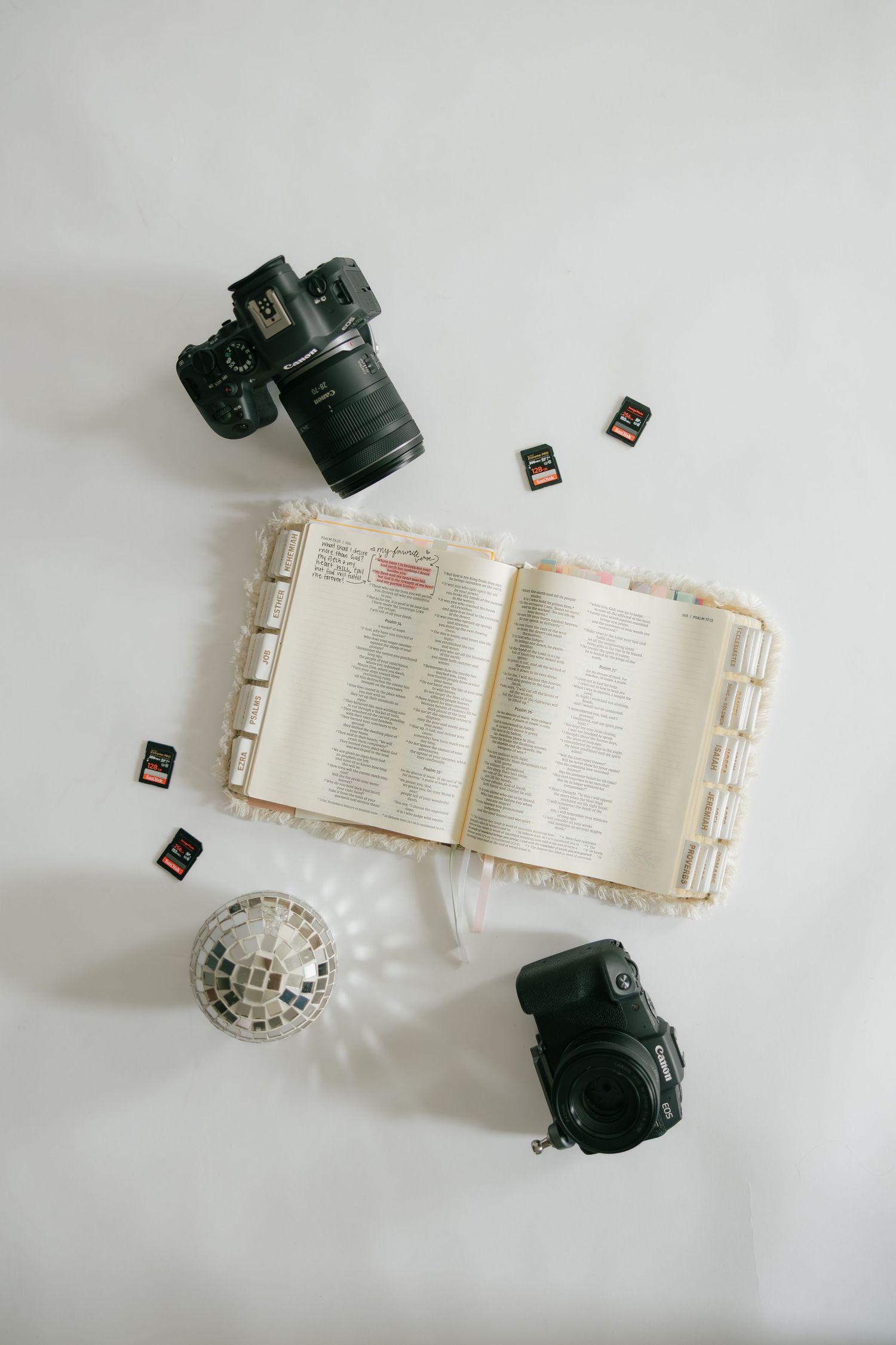 Flat lay of open Bible, DSLR cameras, mini disco ball, and SD cards on white background.