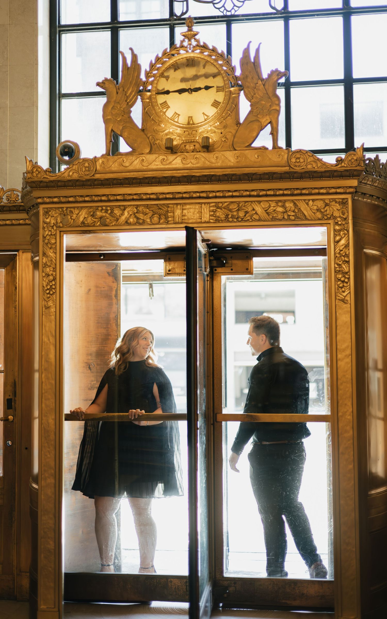 Couple standing in ornate golden revolving door entrance with decorative clock above in elegant historic building.