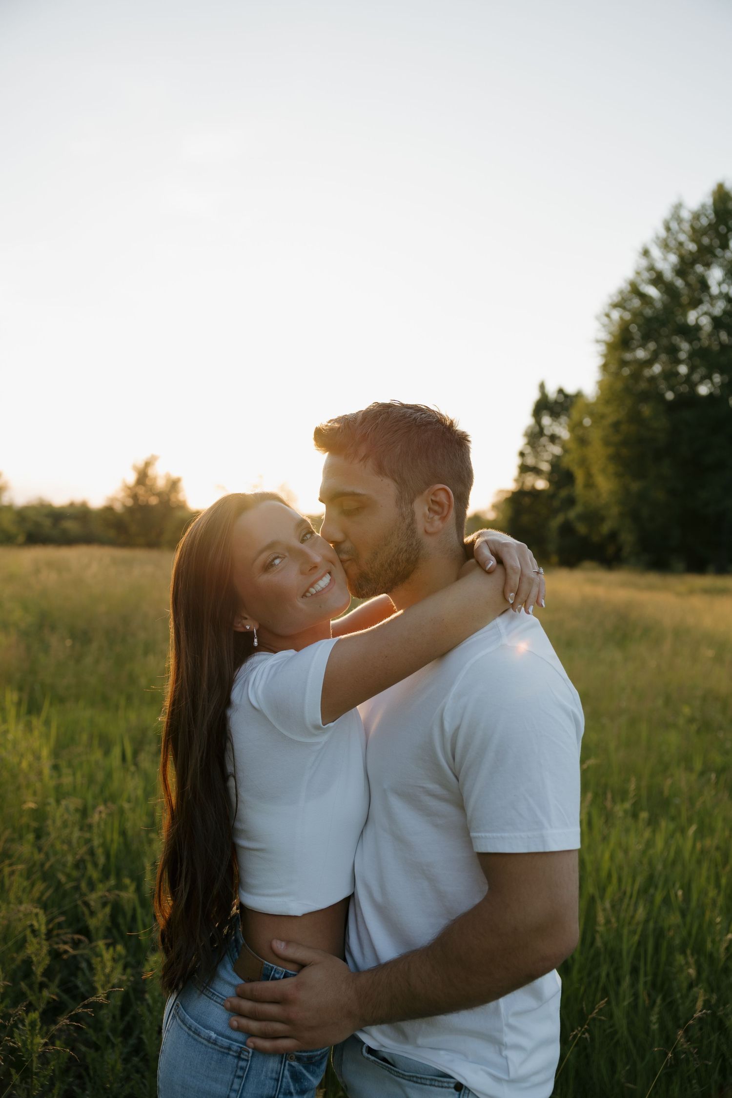 A couple embraces in a sunlit green field at golden hour, the woman smiling as the man kisses her cheek.