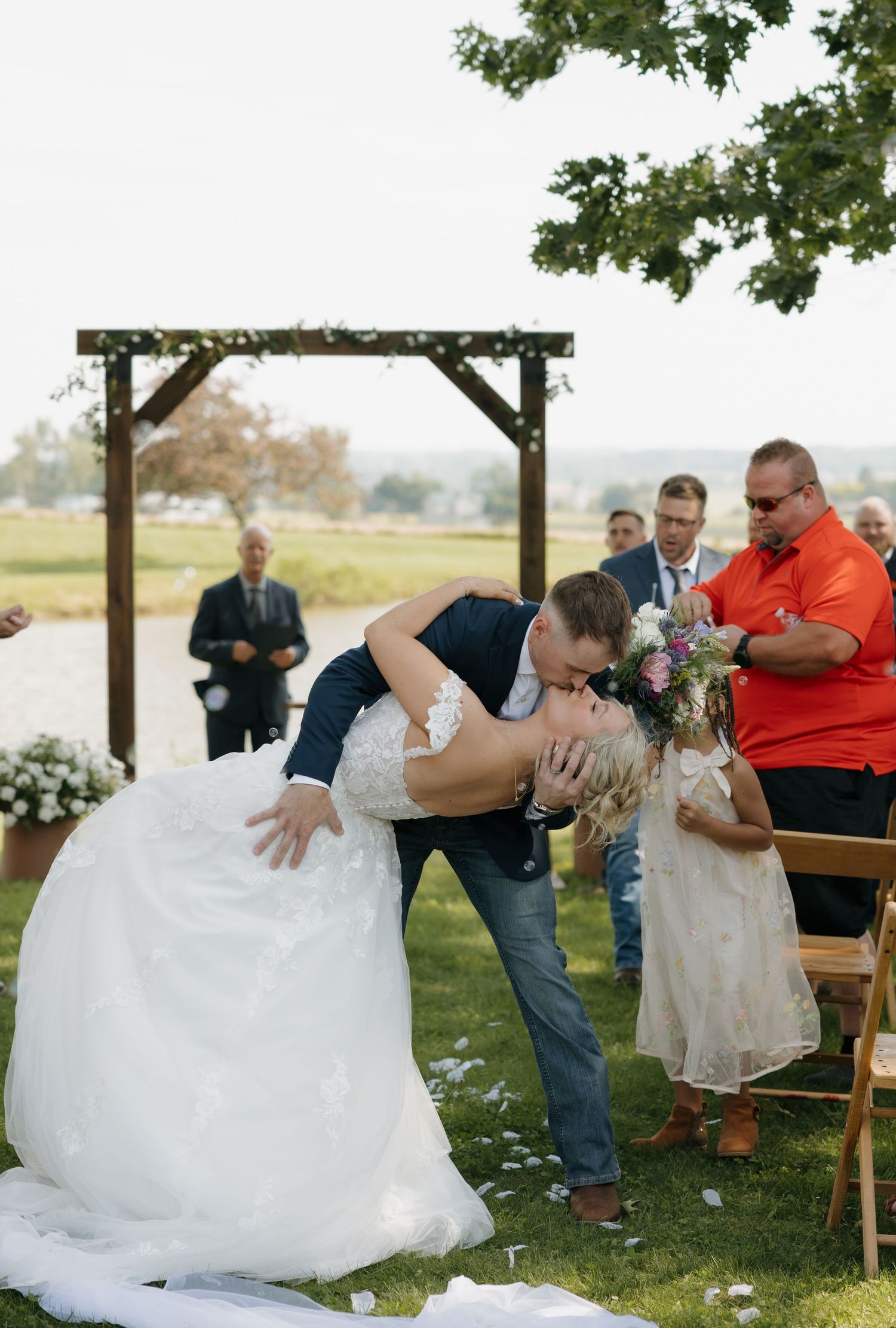 Groom dipping bride for a kiss after outdoor wedding ceremony near wooden arch.