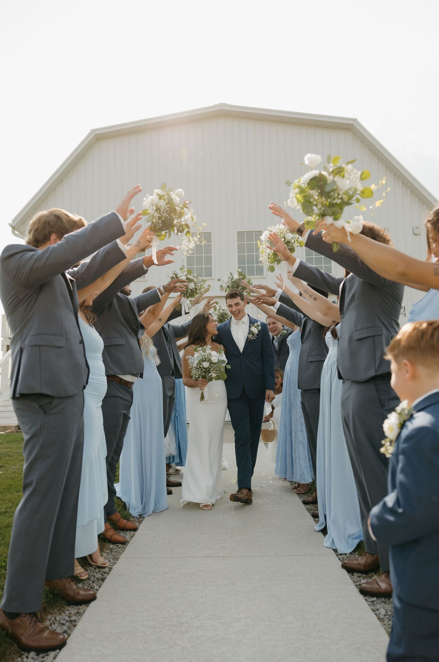 Wedding party tunnel with bridesmaids in blue gowns and groomsmen in gray suits raising bouquets as bride and groom walk through.
