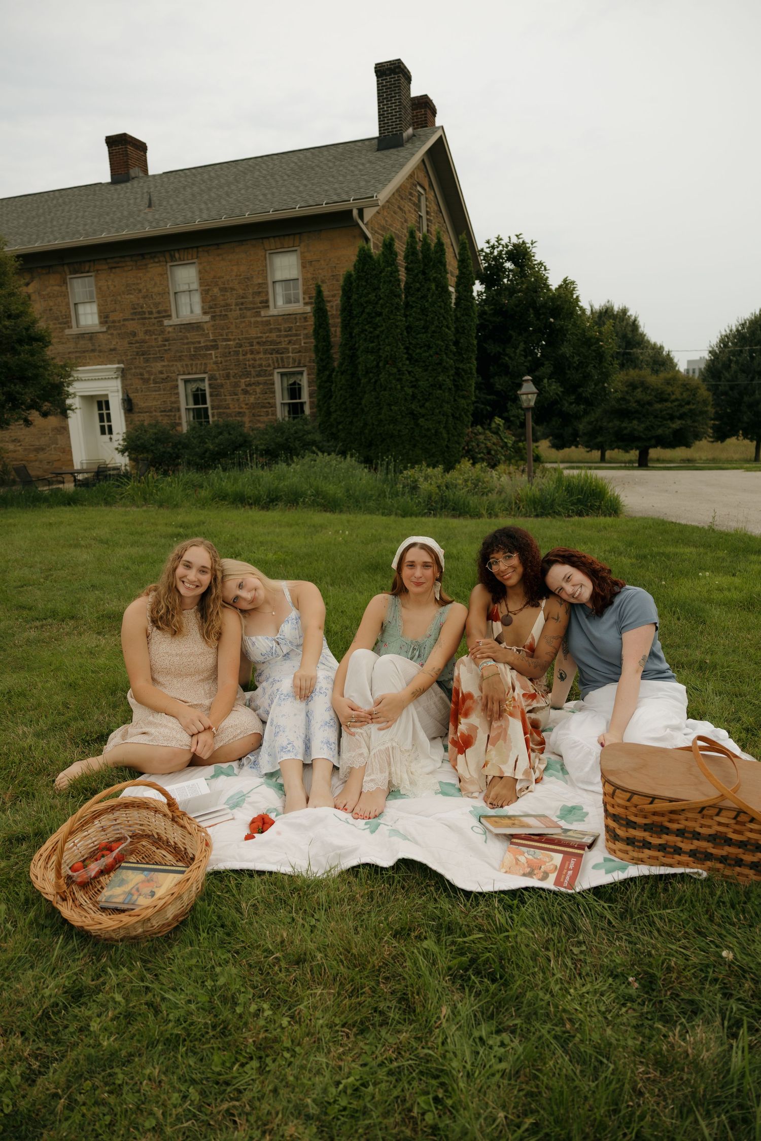 Five women pose together on a picnic blanket with wicker baskets on a lush green lawn near a stone house.