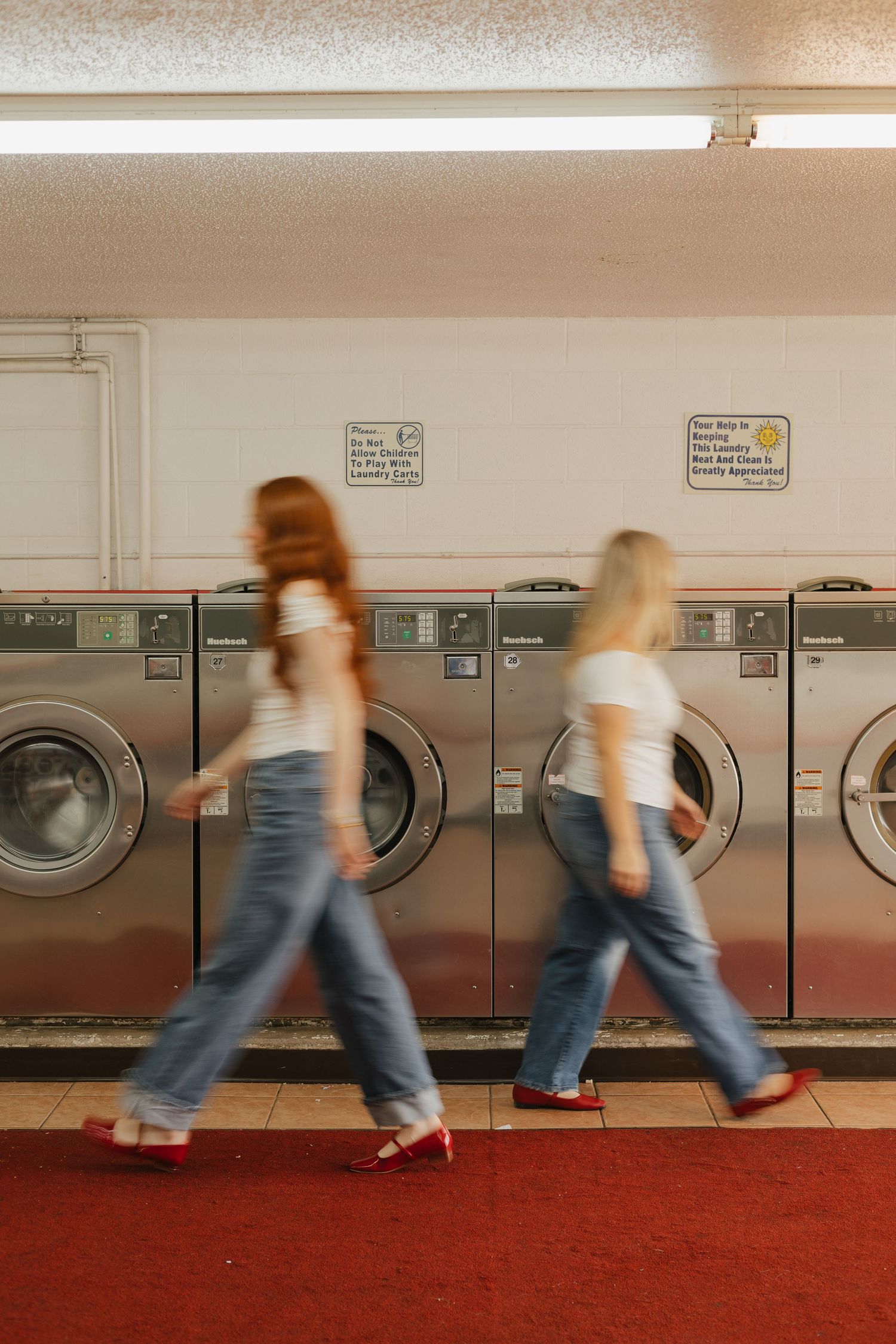 Motion blur captures people walking past a row of washing machines in a laundromat with red carpeting.