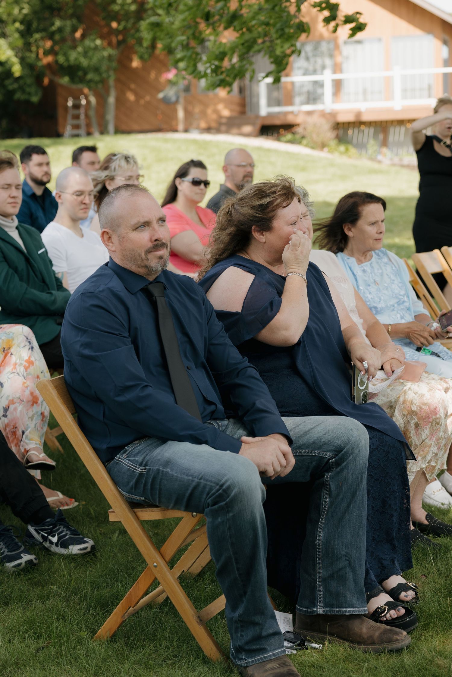 Guests seated outdoors at a wedding ceremony, with a man in navy and a woman wiping tears in the front row.