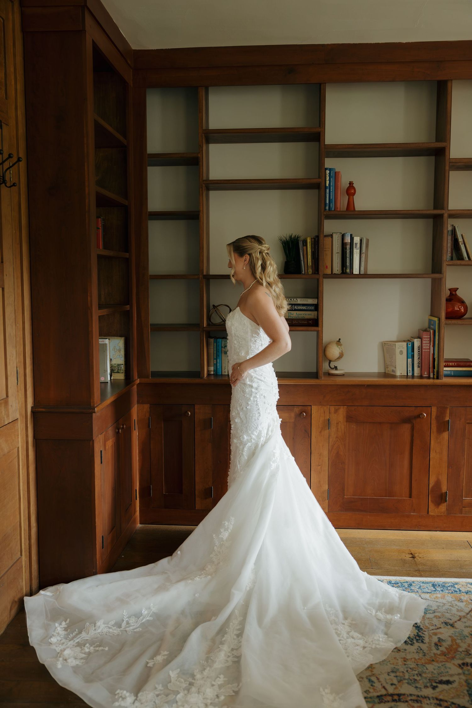 Bride in a strapless lace mermaid wedding gown with train stands in an elegant wood-paneled library.
