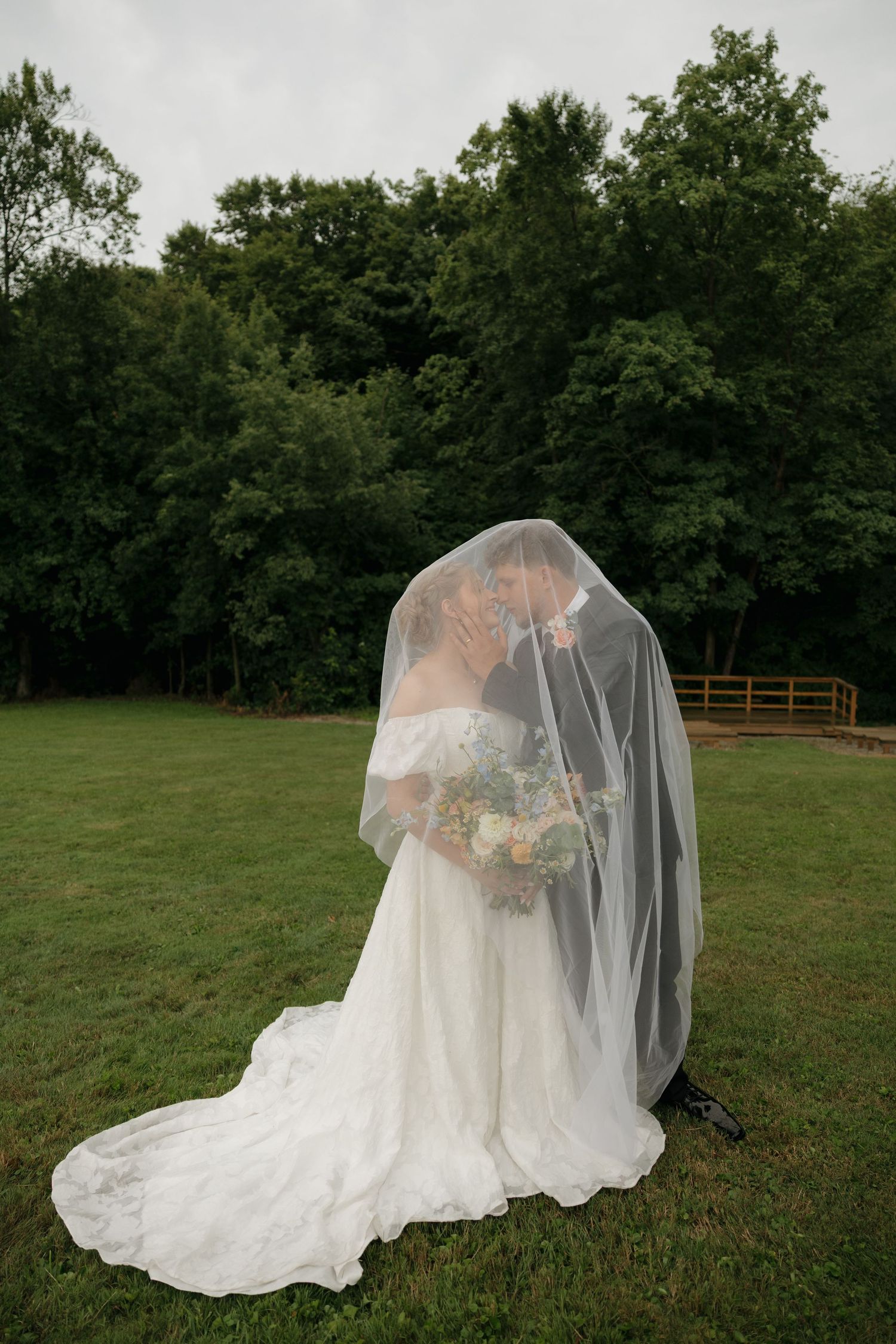 Bride and groom share a kiss under flowing veil on green lawn.