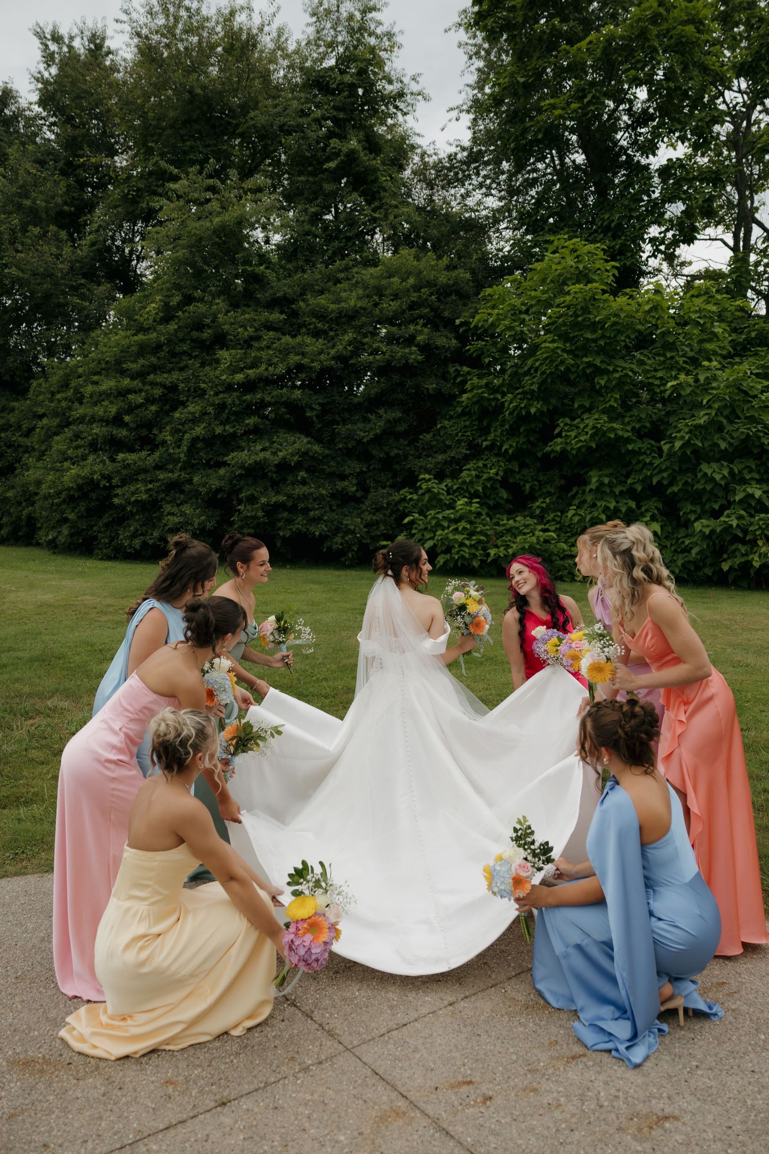 Bride in white gown surrounded by colorful bridesmaids holding bouquets, arranging her train outdoors in a lush green park.