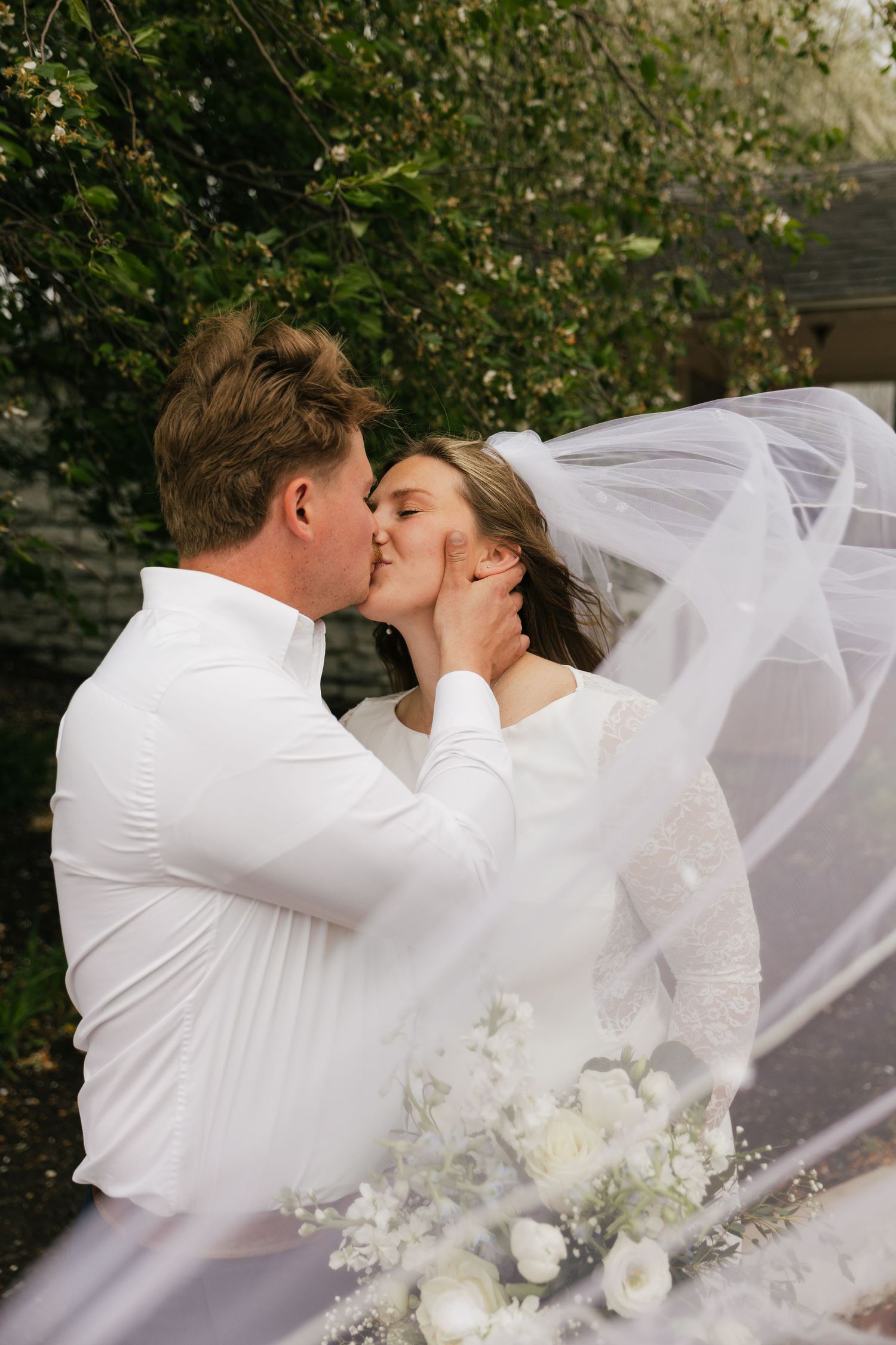 Bride and groom share a romantic kiss outdoors, with her white veil flowing in the wind and a bouquet of white flowers.