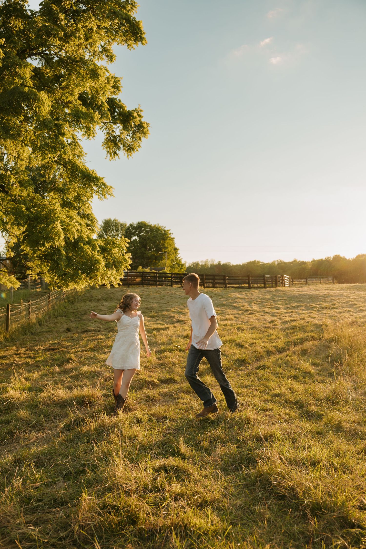 Couple dancing playfully in a sunlit green field at golden hour, with trees and a wooden fence in the background.