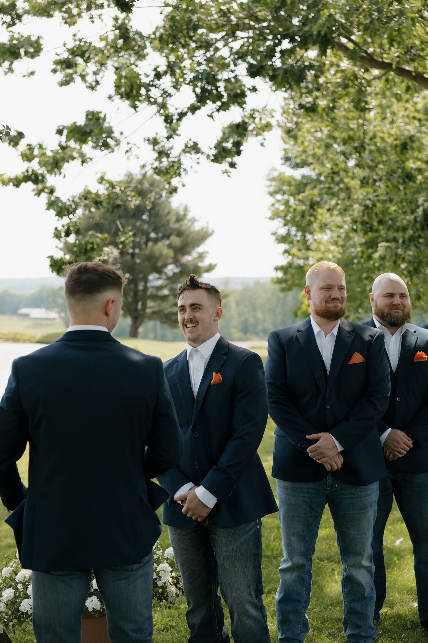 Groom smiling at groomsmen in navy suits with orange pocket squares at outdoor wedding.