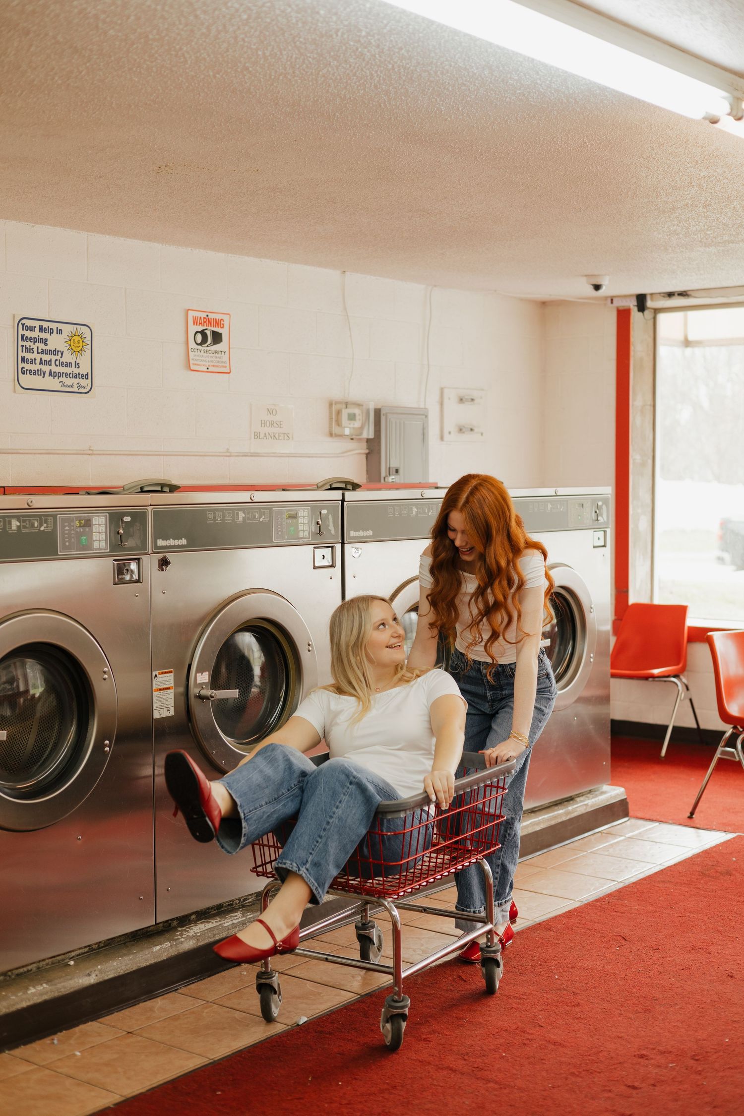 Two people relax in chairs at a retro laundromat with red carpet and stainless steel washing machines.