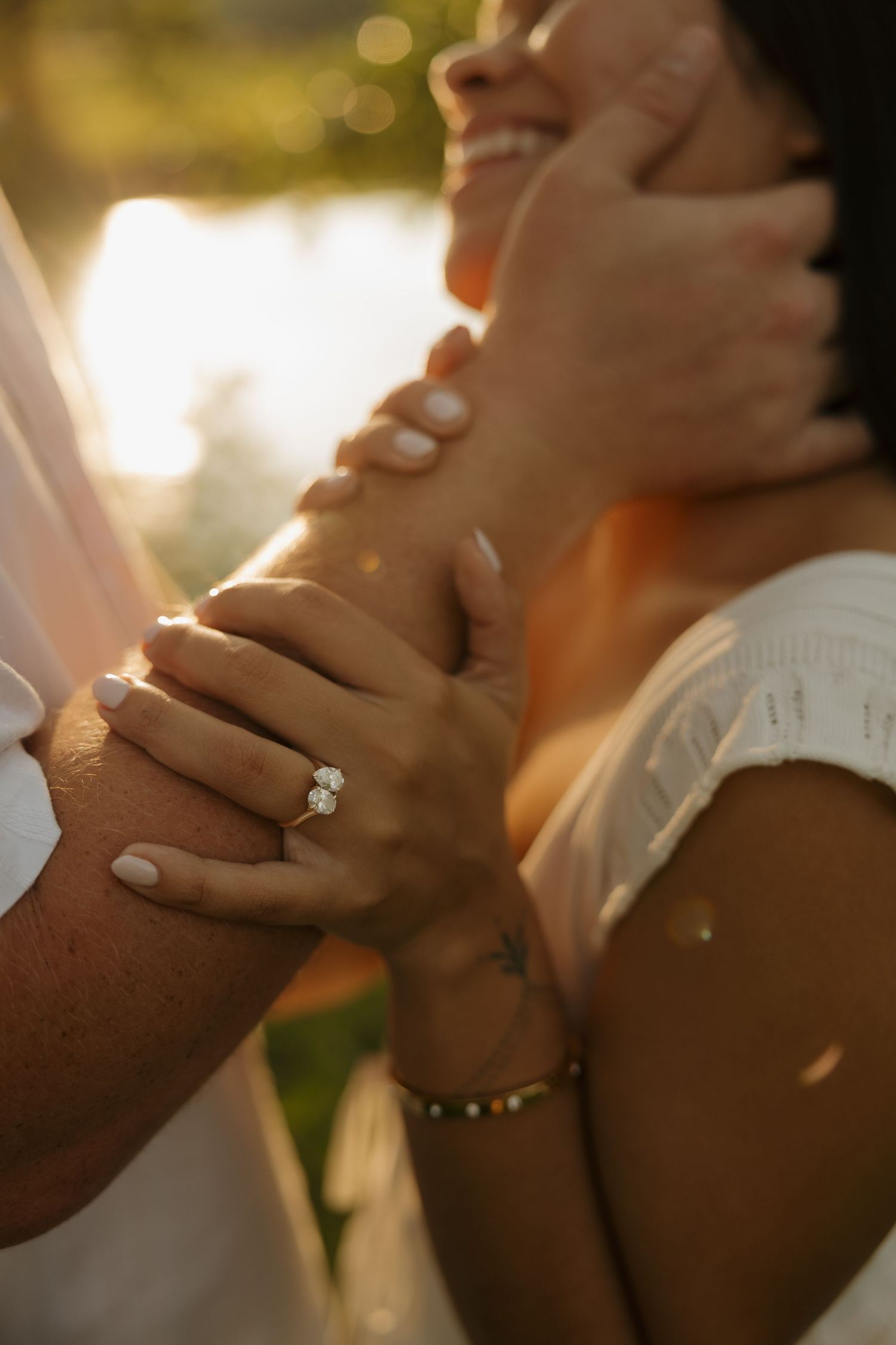 Close-up of a woman's hand wearing a diamond engagement ring, held tenderly by her partner in golden sunset light.