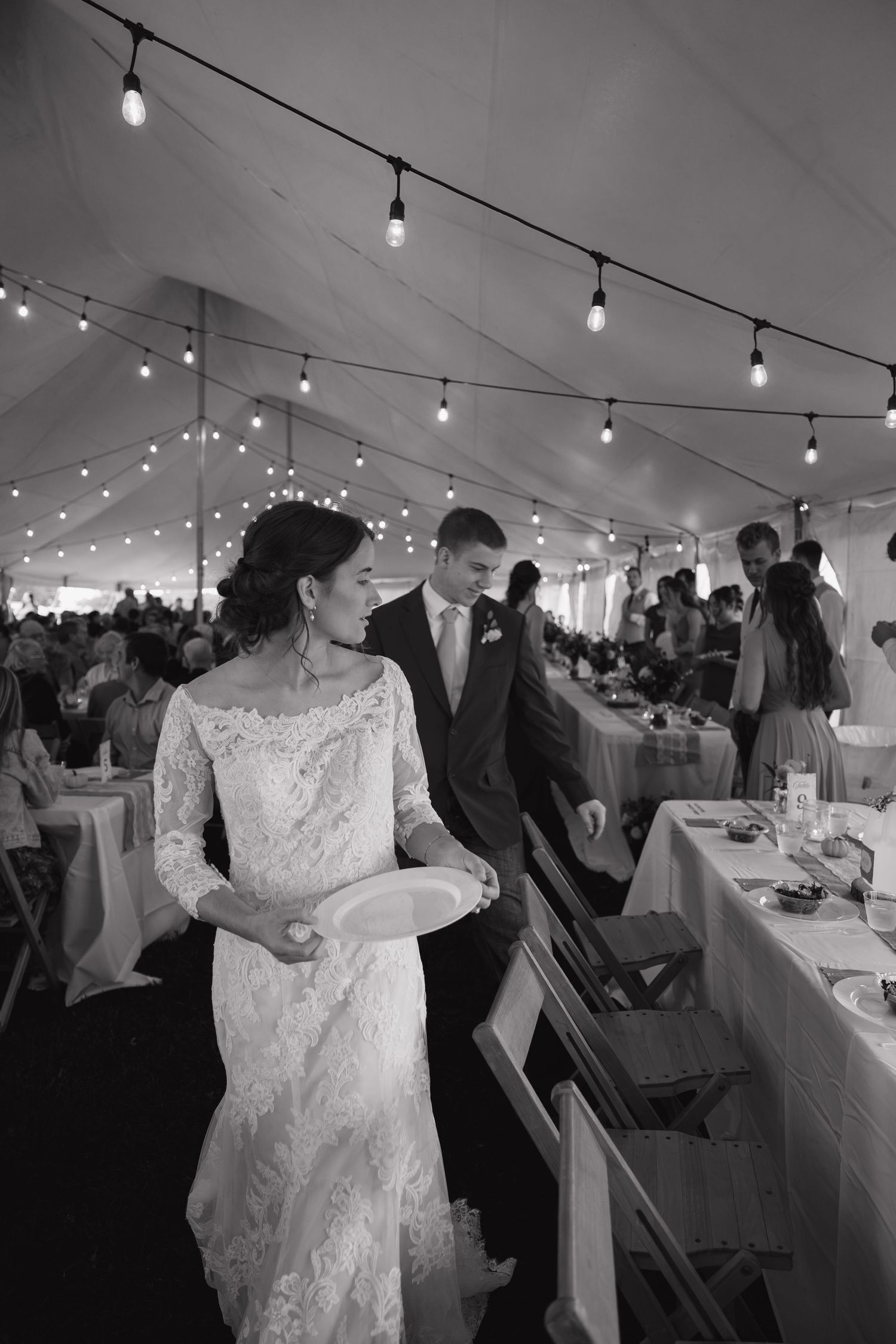 Bride in a lace wedding gown holds a plate beside the groom as they walk through a tent reception with string lights and guests.