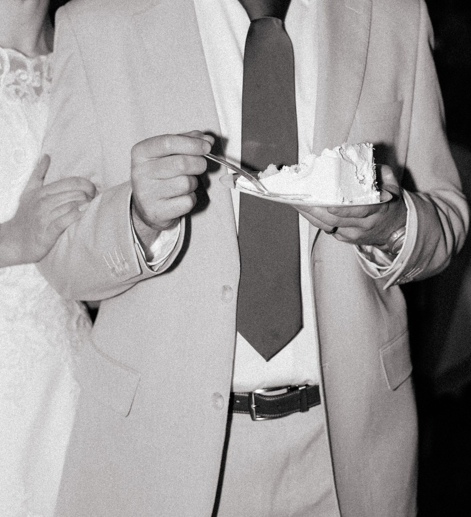 Black and white photo of a groom in a suit holding a slice of wedding cake with a fork.