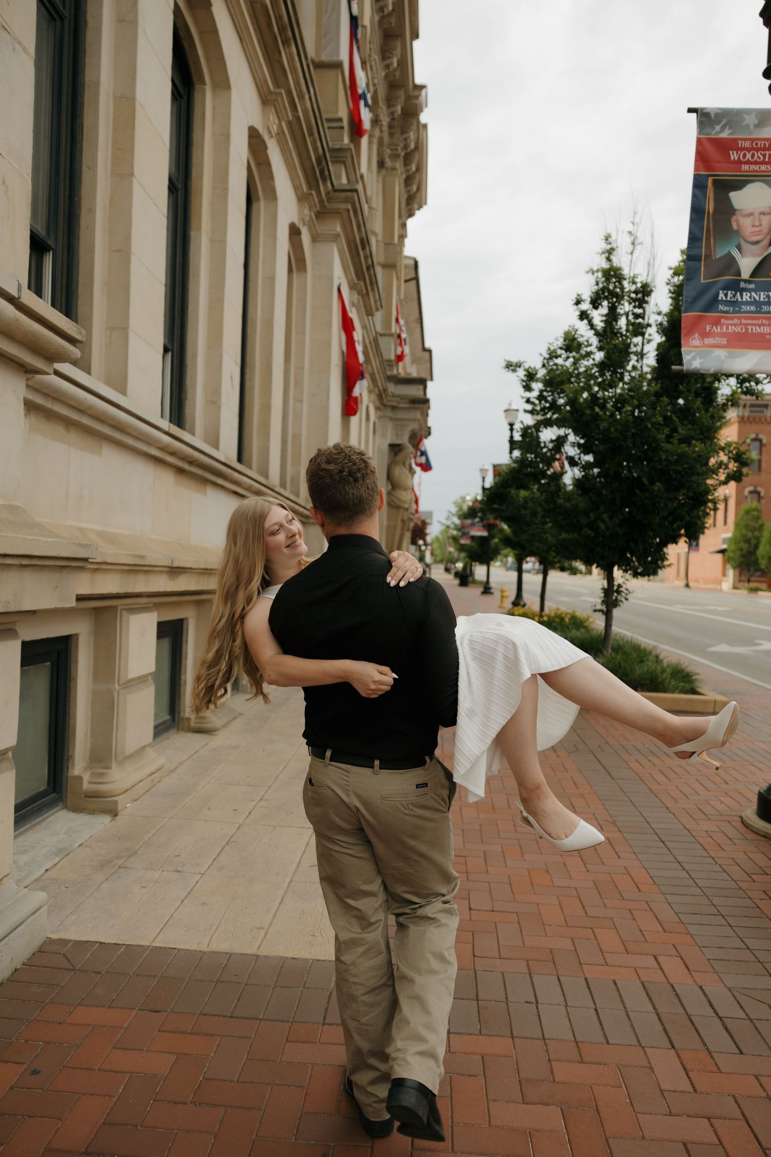 A man in a black shirt carries a woman in a white dress on a brick sidewalk outside a historic building.
