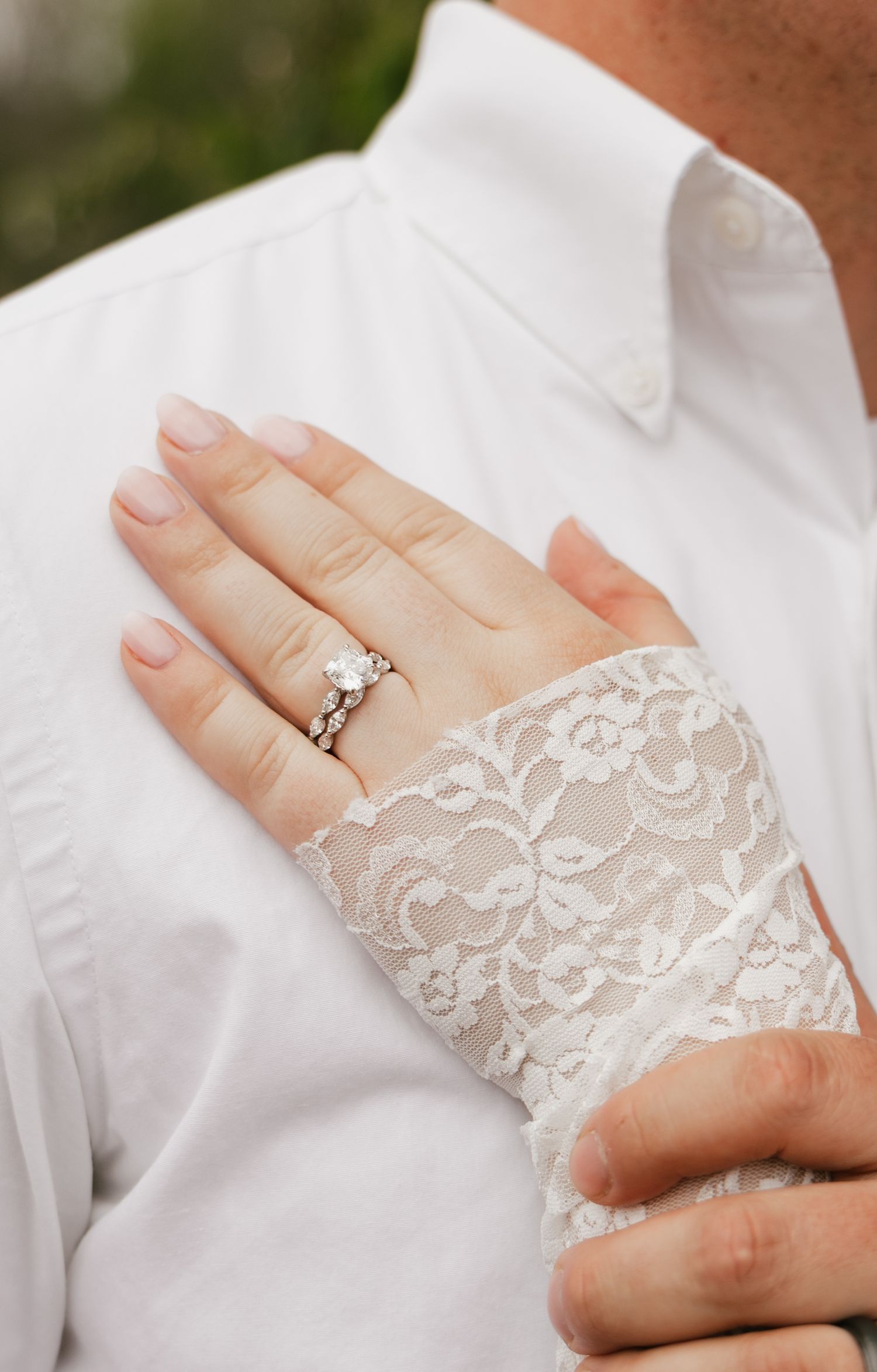 Close-up of a woman's hand with diamond wedding ring set, resting on a man's white shirt, wearing lace sleeve.